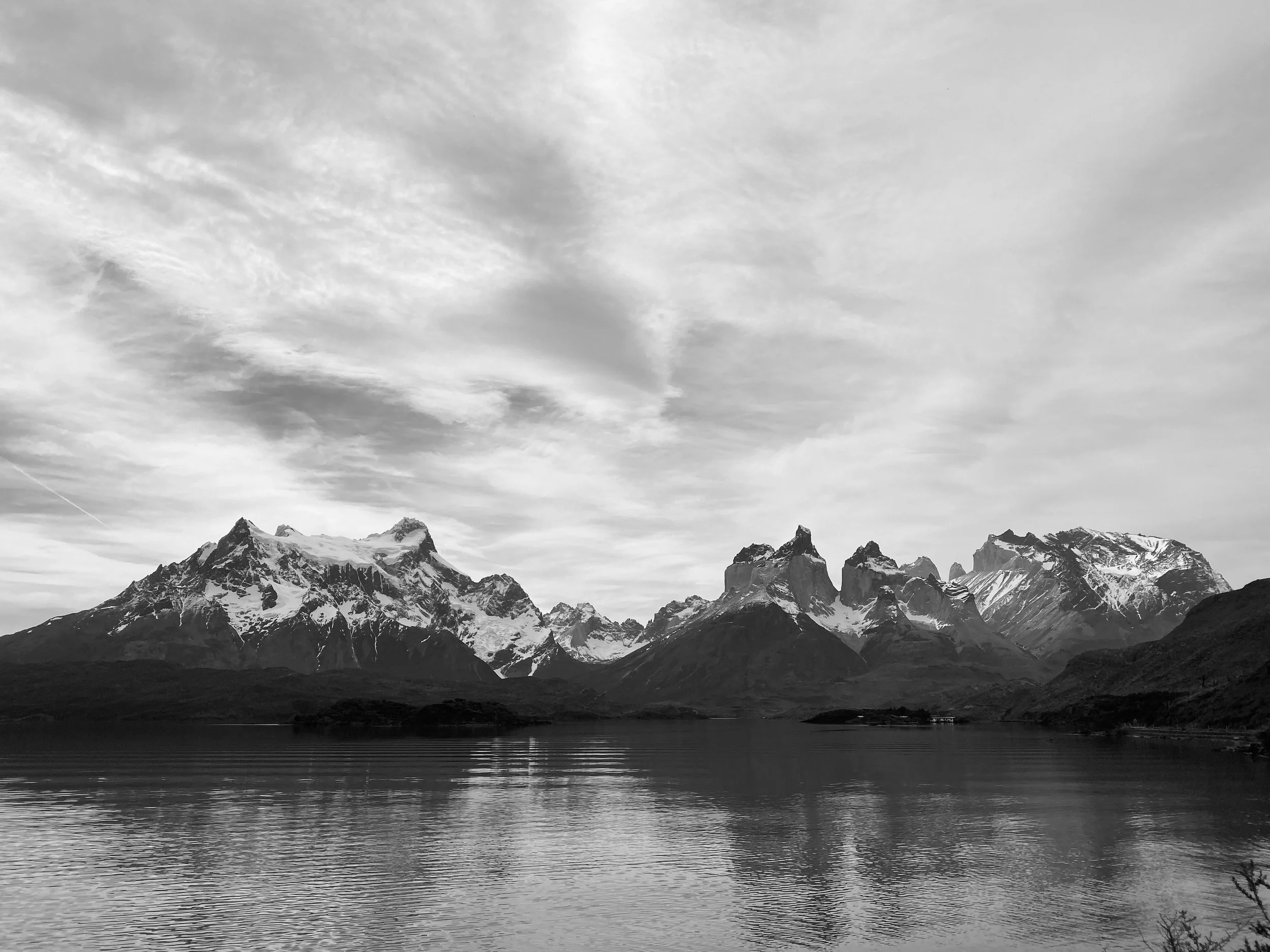Black and white photograph of snow-capped mountains reflecting in a calm lake beneath a cloudy sky.