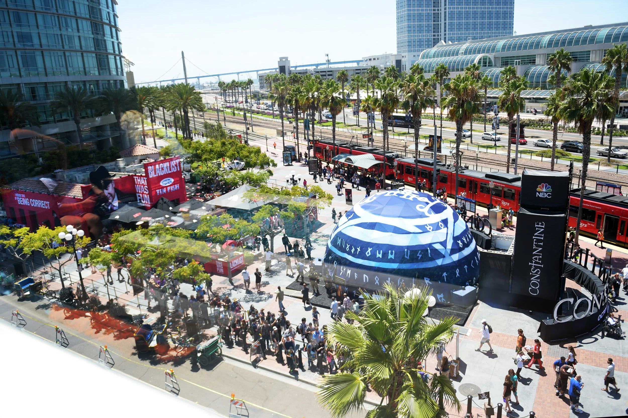 People gather outside a convention center with banners and tents, red train at a stop, and modern glass buildings, palm trees, and a bridge in the background.