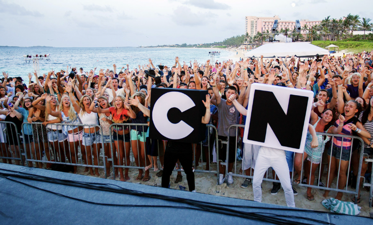 Large crowd of people on the beach holding up large letters 'C' and 'N' during a live event.