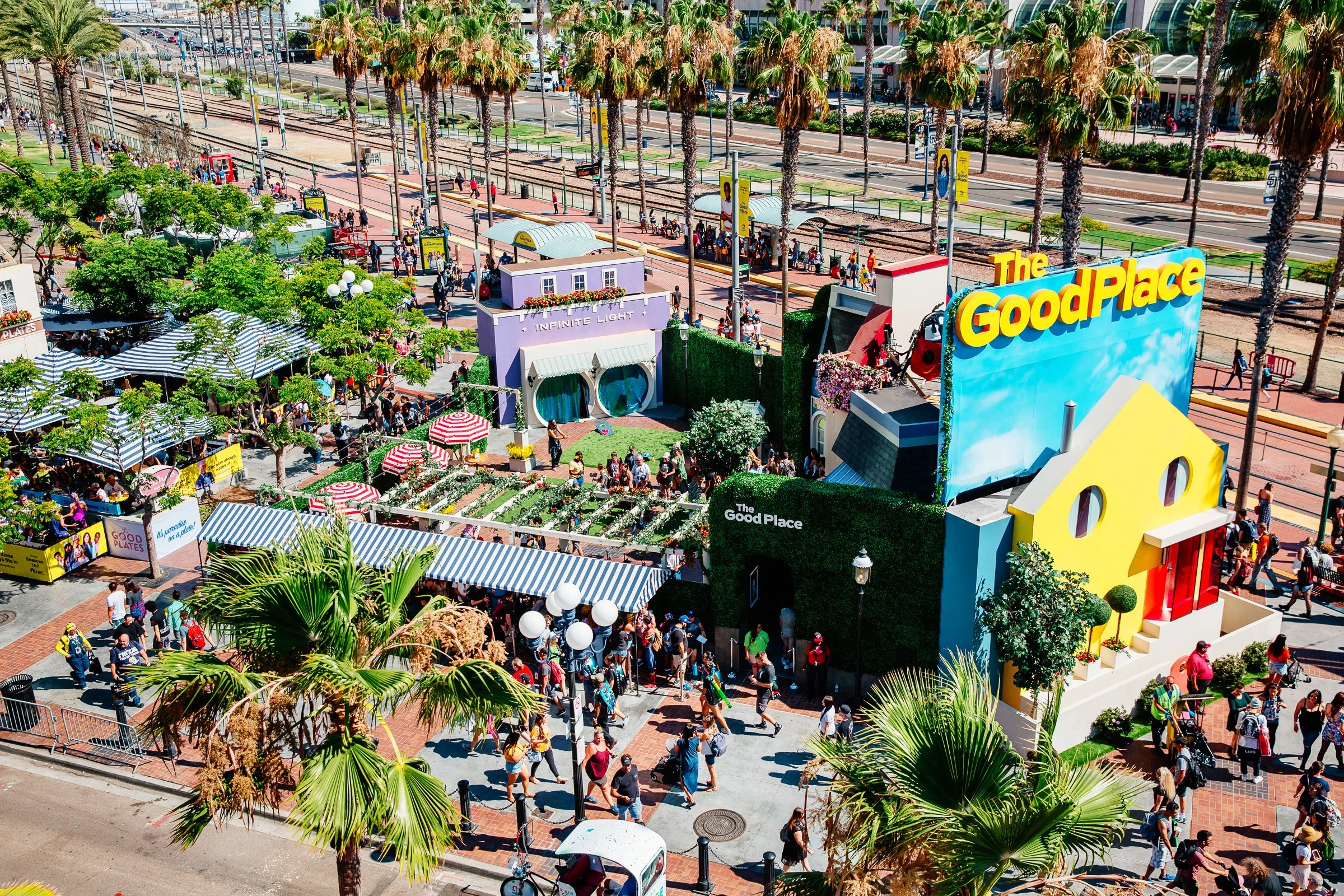 A busy outdoor shopping and entertainment area with palm trees, colorful buildings, and many people walking and waiting in lines.