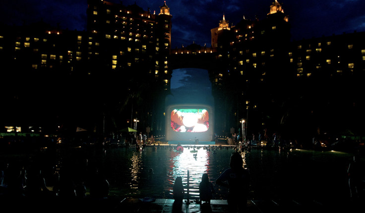 Night view of an outdoor swimming pool surrounded by tall illuminated buildings, with a large screen showing a cartoon character, and people sitting and swimming in the pool.