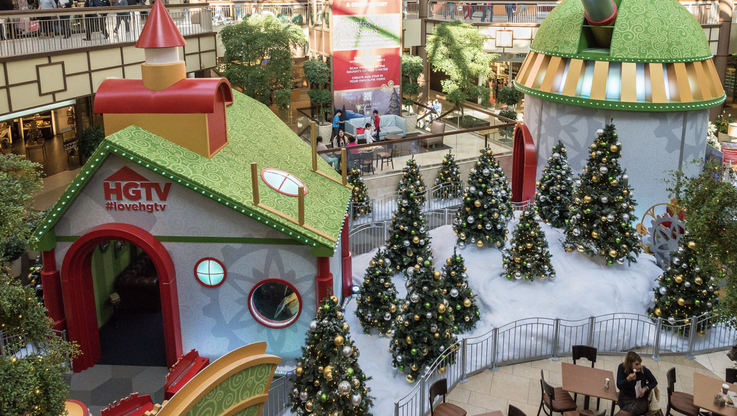 Christmas display inside a shopping mall, featuring various decorated Christmas trees with gold and silver ornaments, surrounded by green trees and holiday decorations. Two colorful, whimsical house-like structures with green, red, and yellow roofs are visible, along with a sign for HGTV.