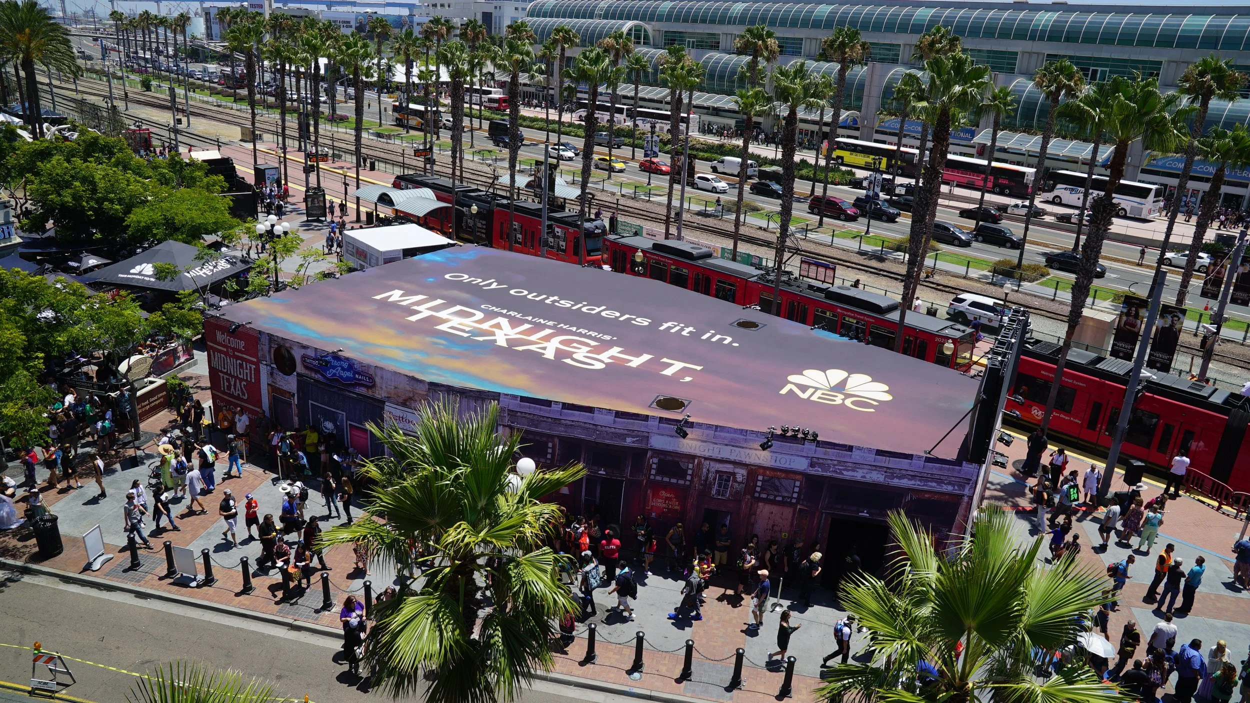 Aerial view of a busy outdoor area with people walking and lining up, palm trees, a train, and a large billboard with NBC advertisement in downtown Dallas, Texas.