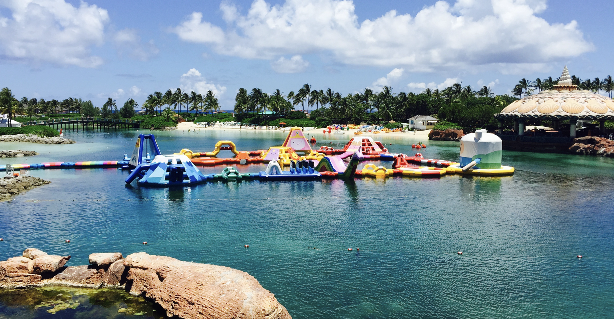 Colorful inflatable water playground on a tropical beach with palm trees, sandy shore, and blue sky.