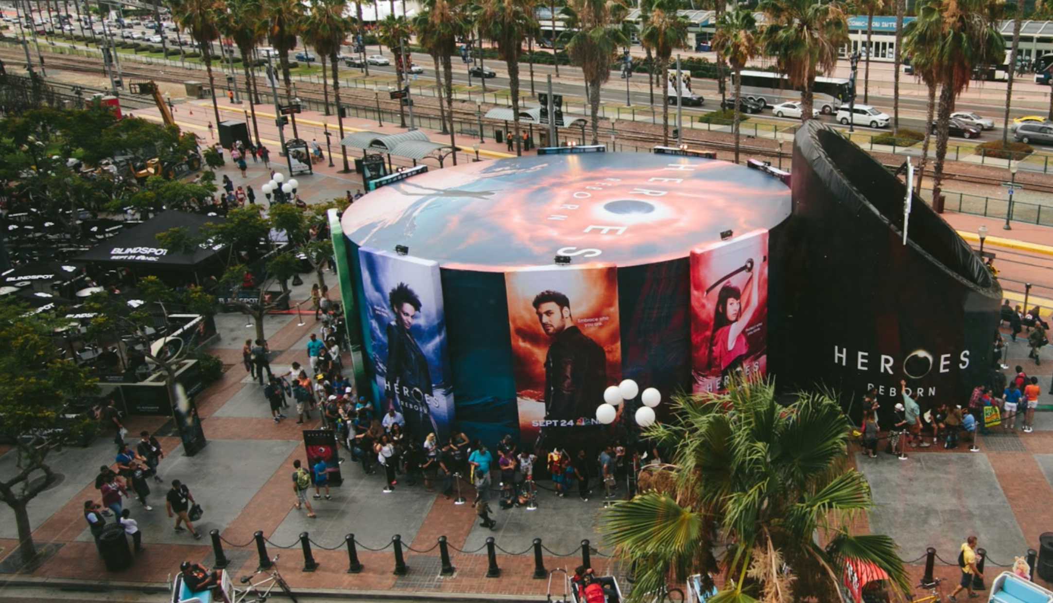 Crowded outdoor urban event space with large circular promotional display featuring movie posters, surrounded by people, trees, and storefronts along a busy street.