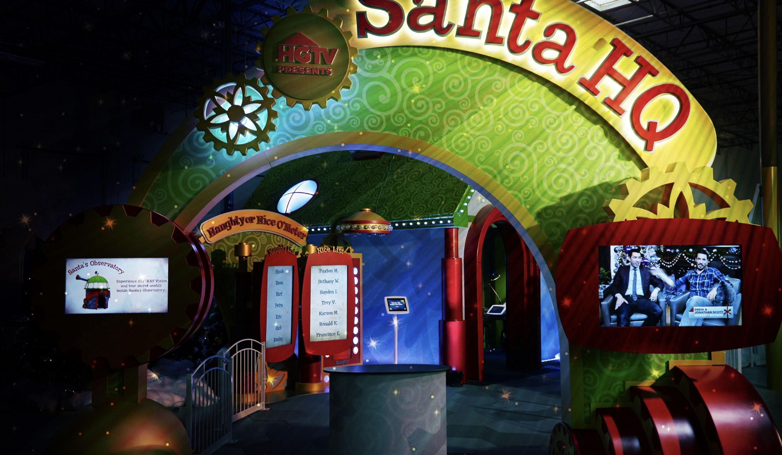 Colorful amusement or entertainment setup with gears and decorative elements, featuring a screen with two men seated, and a sign for Naughty or Nice Meter with list of names.