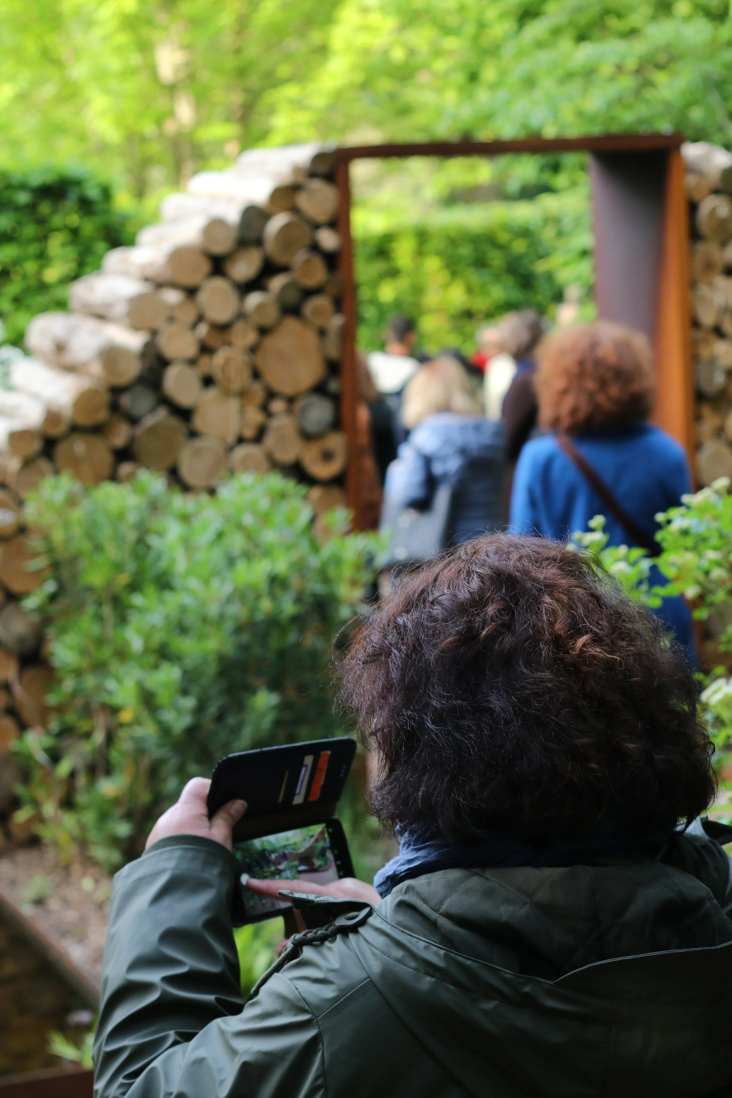 People gathered outdoors near a wood pile, with one person taking a photo of the group against a natural green background.