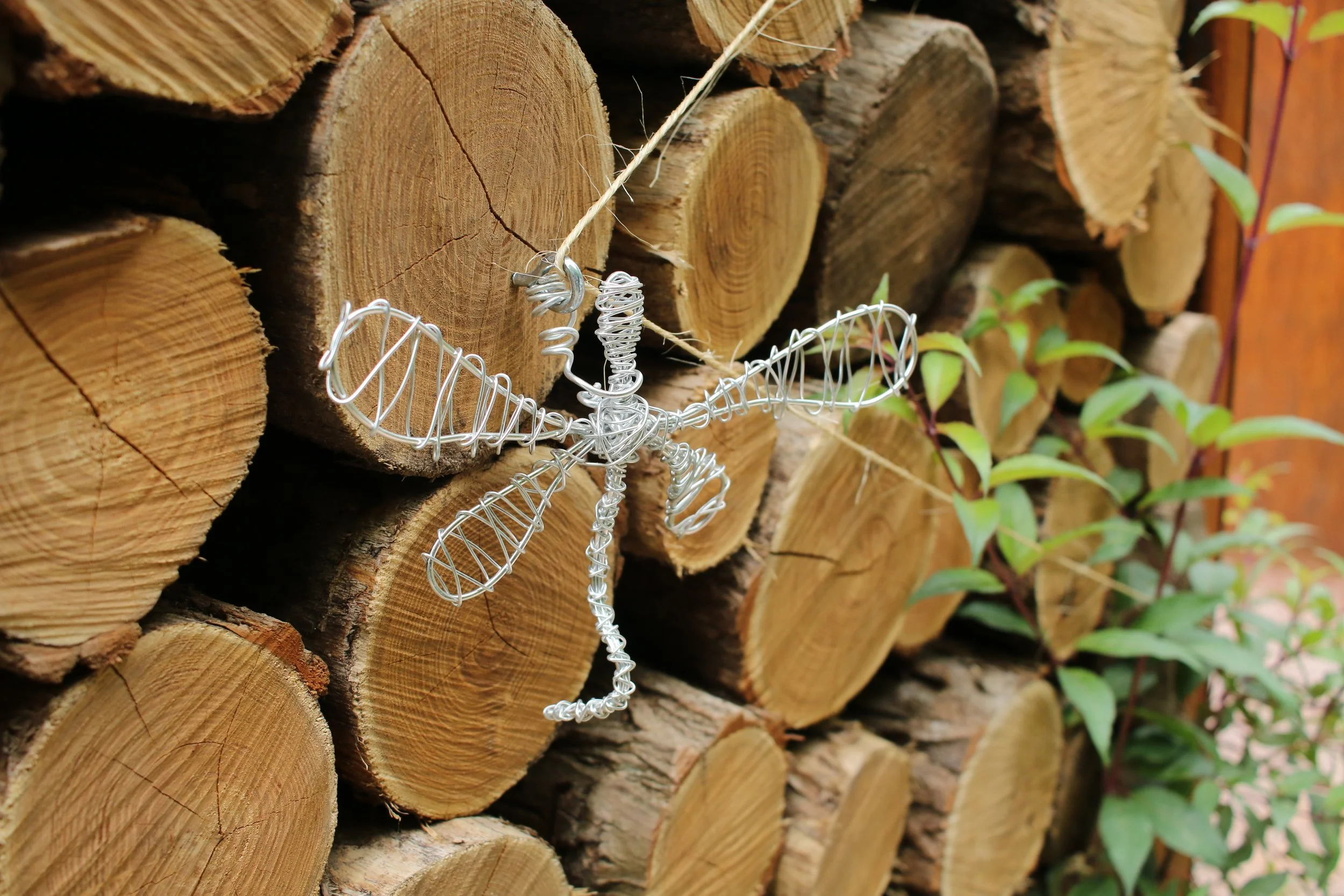 A wire sculpture of a dragonfly mounted on a stack of cut logs with green leaves nearby.