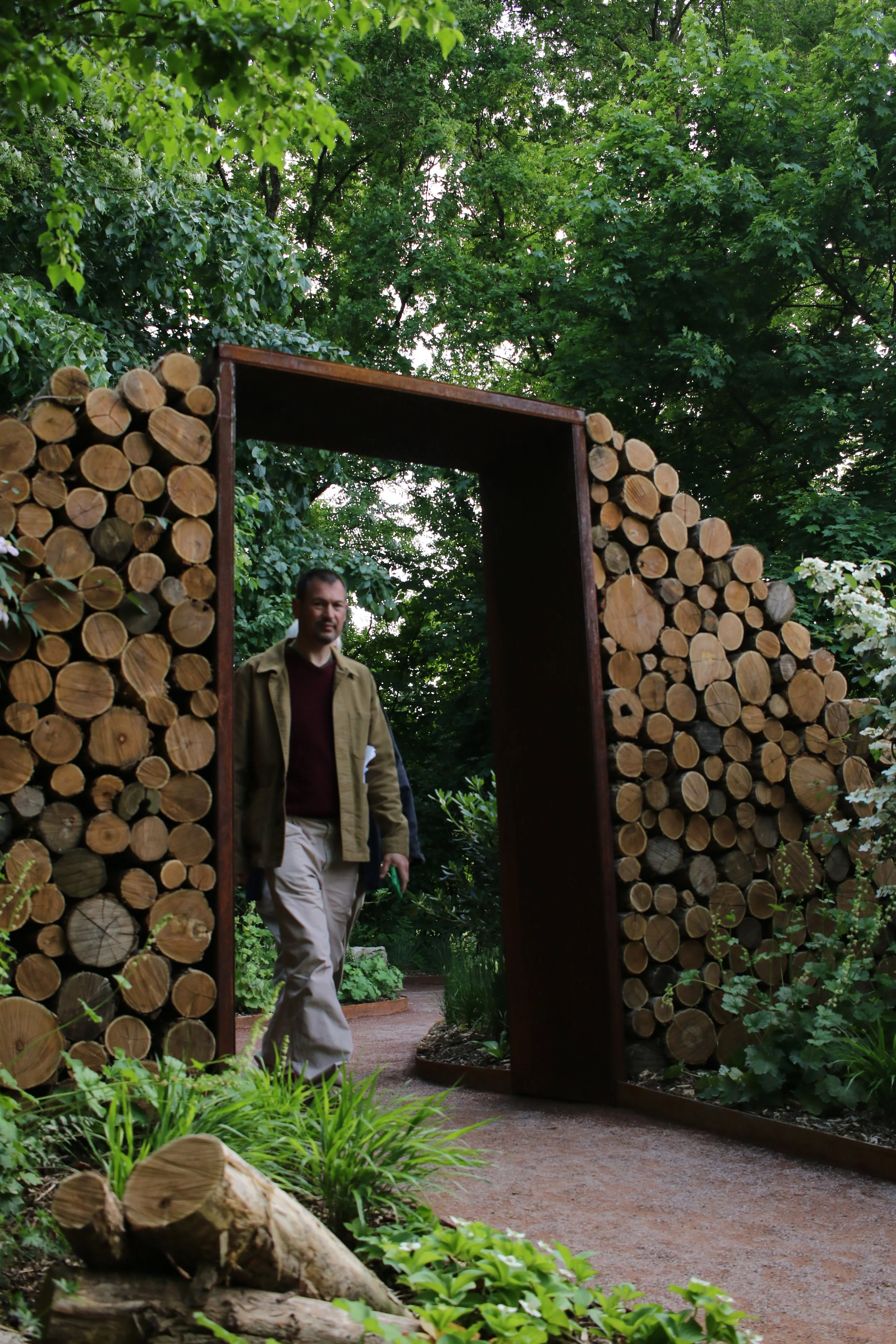 A man walking through a garden pathway framed by a modern arch and wall made of stacked logs, surrounded by lush greenery.