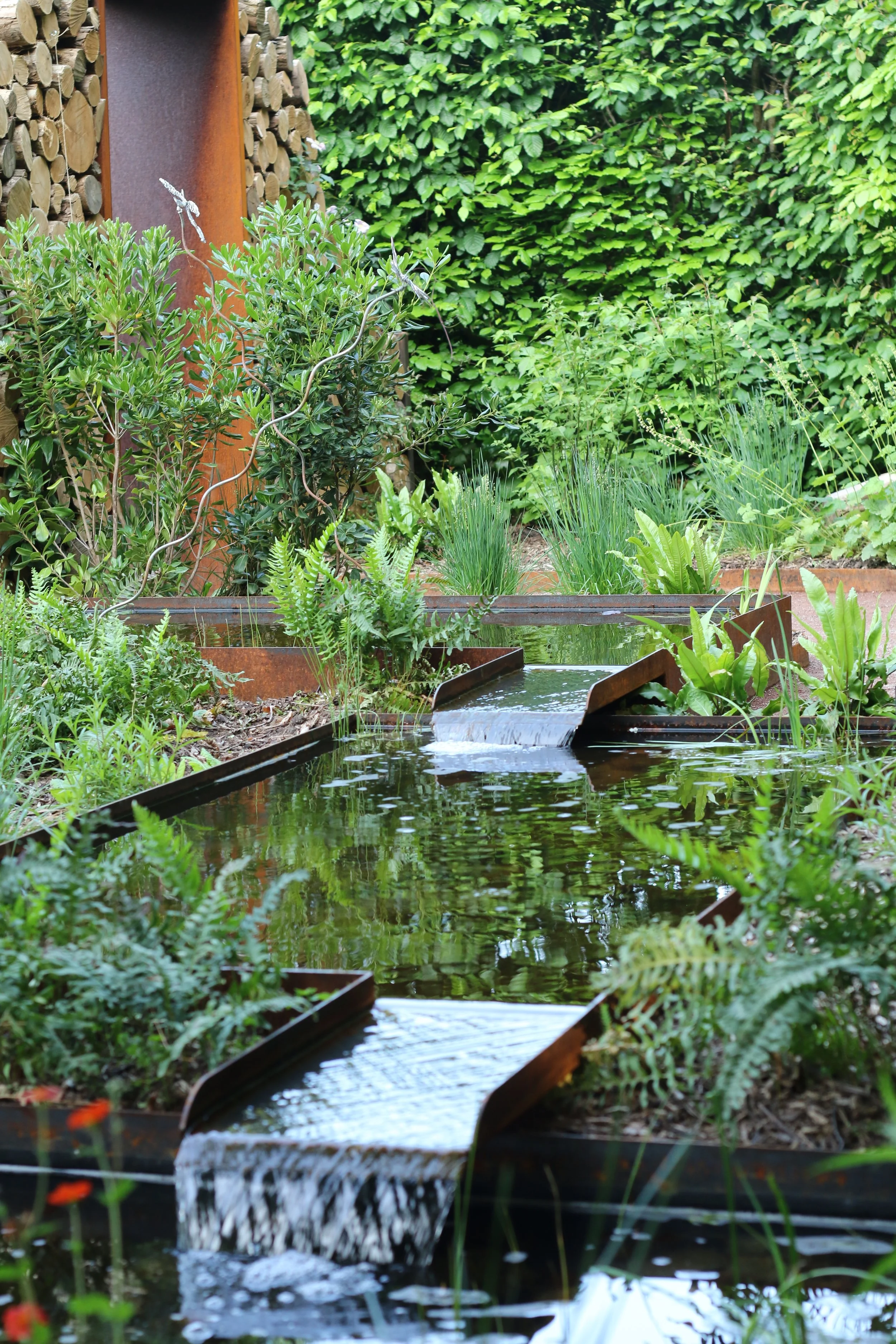 A garden with a water feature flowing through a series of stepped metal channels, surrounded by lush green plants and bushes.