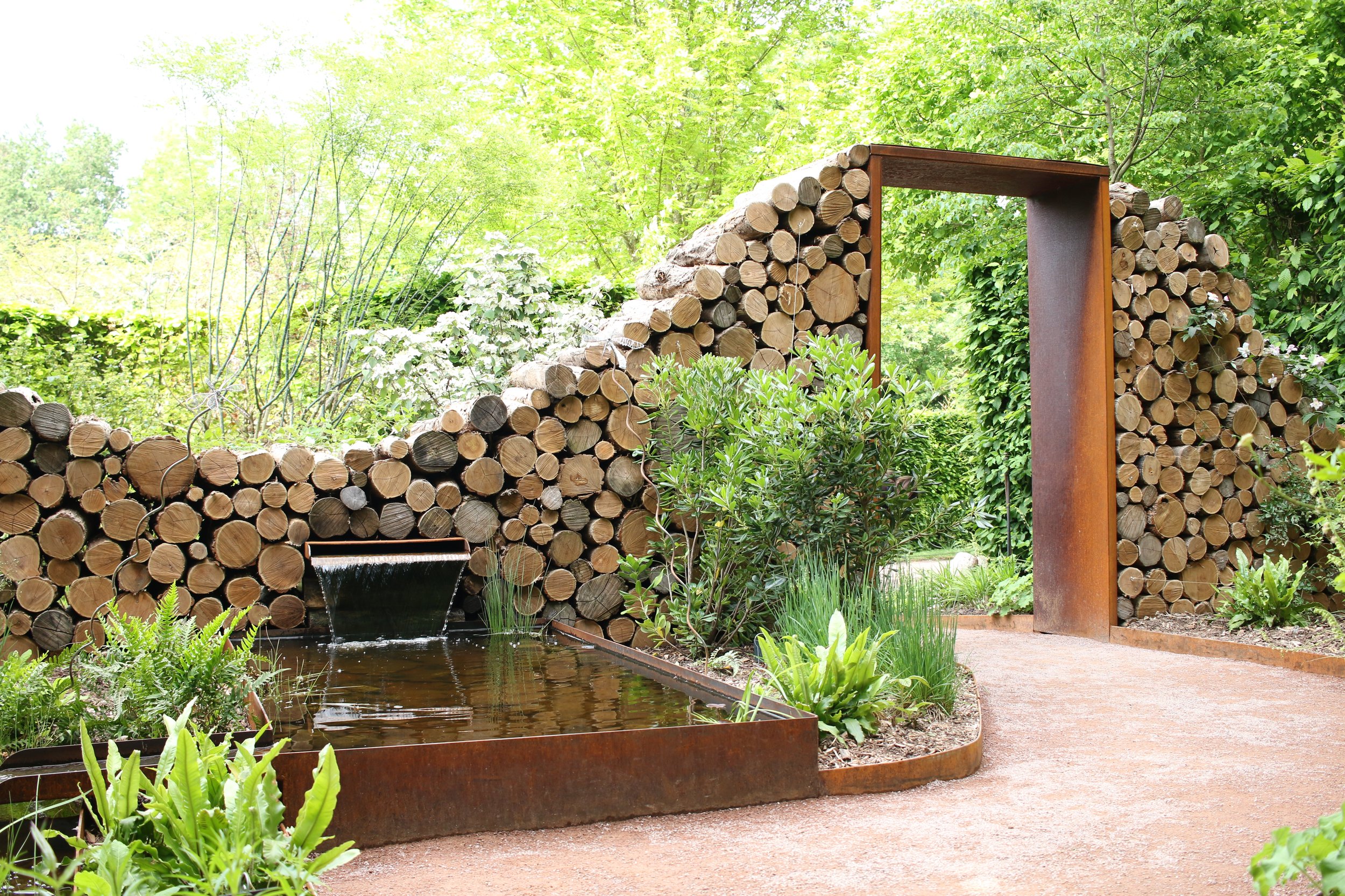 A garden scene with a water feature, stacked logs wall, and an arched wooden and rust metal door in a lush green setting.