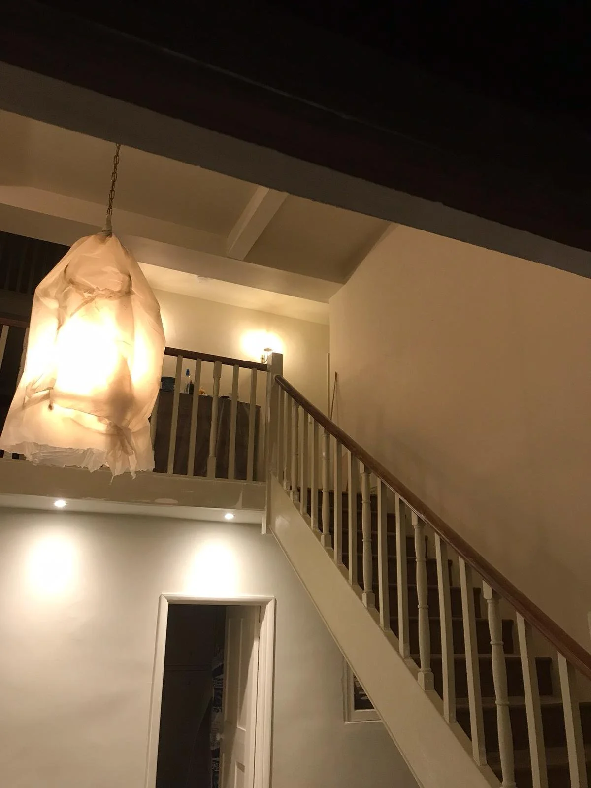 View of an indoor staircase with wooden handrail, leading to a second floor, with a large hanging light fixture and wall-mounted light.