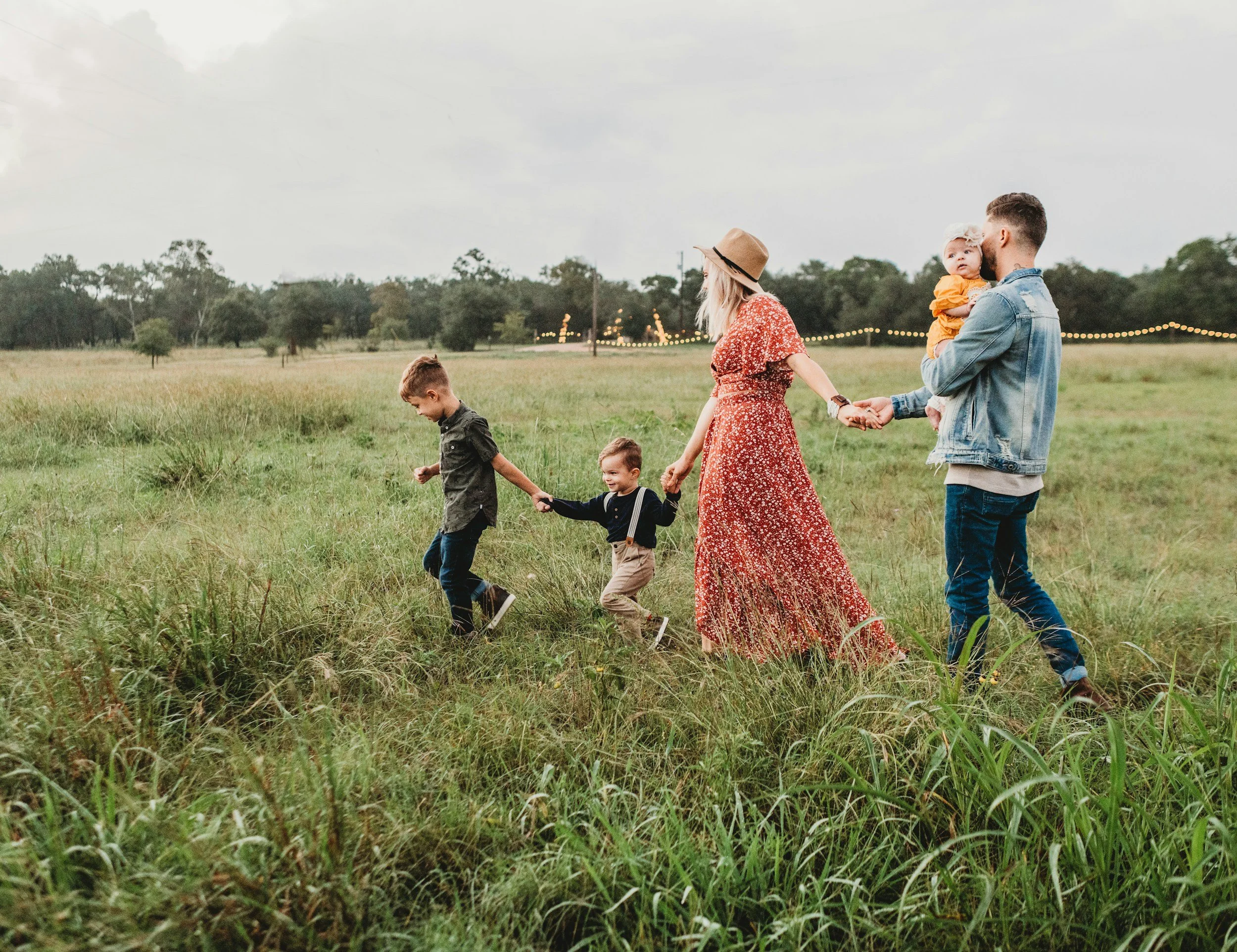 Family walking through a grassy field, holding hands, with a boy leading and a man carrying a young girl, during evening in an open park.