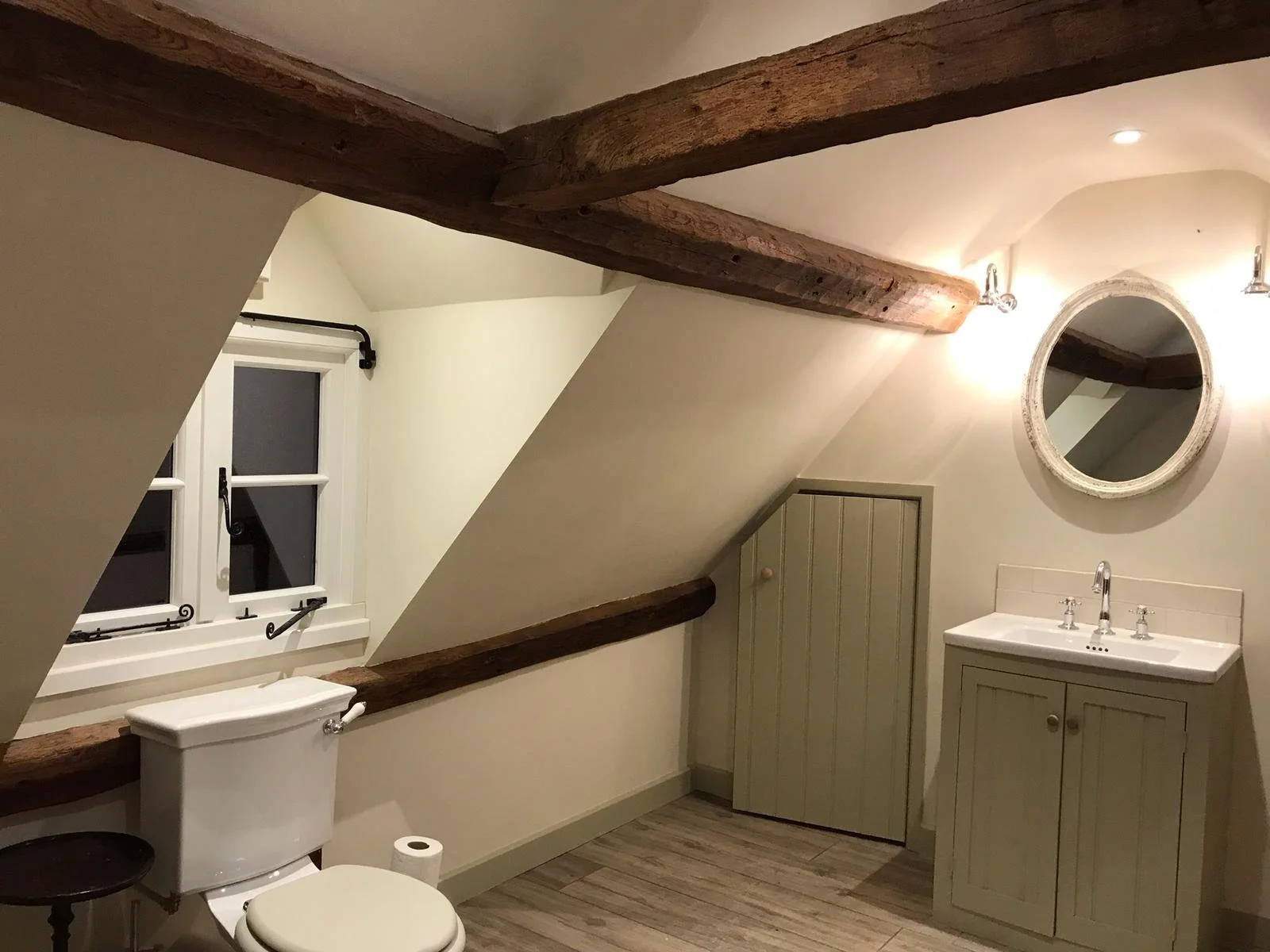A small bathroom with sloped ceiling, exposed wooden beams, a white toilet, a window with black curtain rod, a small vanity with a mirror and sink, and wooden flooring.