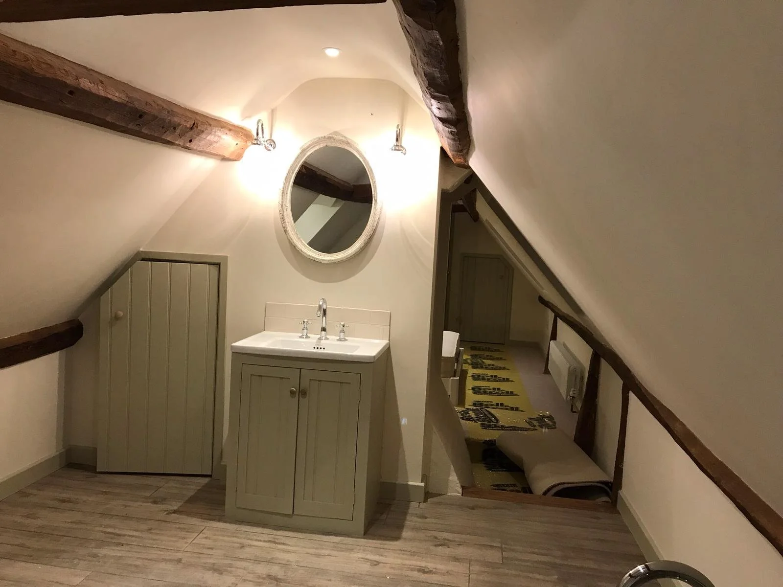 Small bathroom with sloped ceiling, wooden beams, a round mirror, a sink with a cabinet, and a small storage closet. There is a wooden floor and soft lighting.