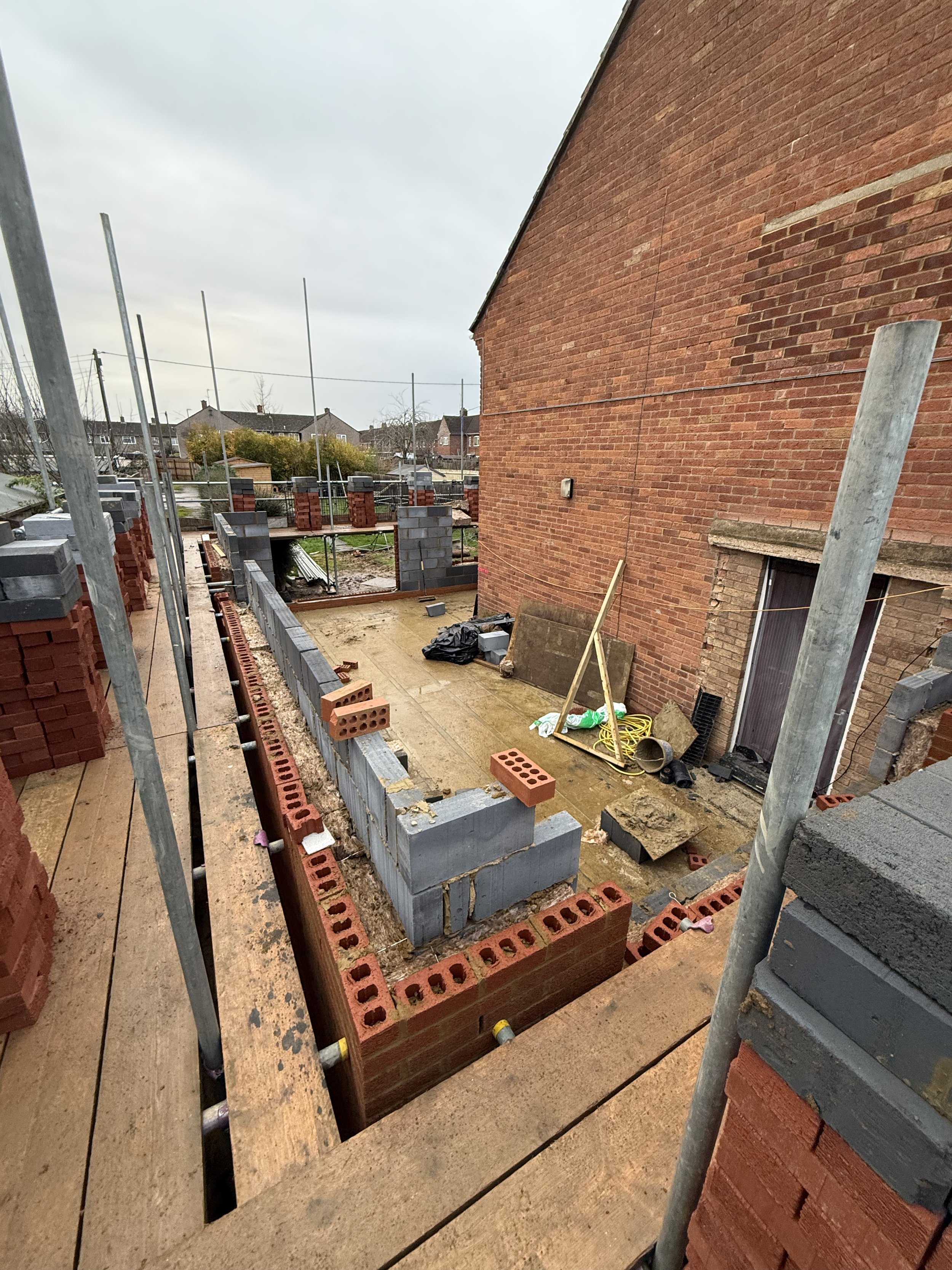 Construction site showing a backyard with partially built brick walls, scaffolding, and construction tools, with neighboring houses in the background.