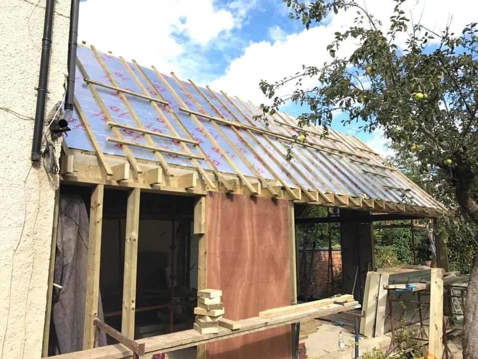 Construction site of a small building or extension with a wooden roof frame in progress, partially covered with roofing underlayment, surrounded by tools, materials, and a nearby tree with green apples.