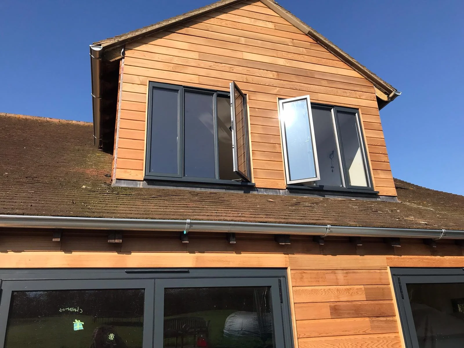A modern house with wooden siding, black window frames, and open dormer windows on a sloped roof, under a clear blue sky.