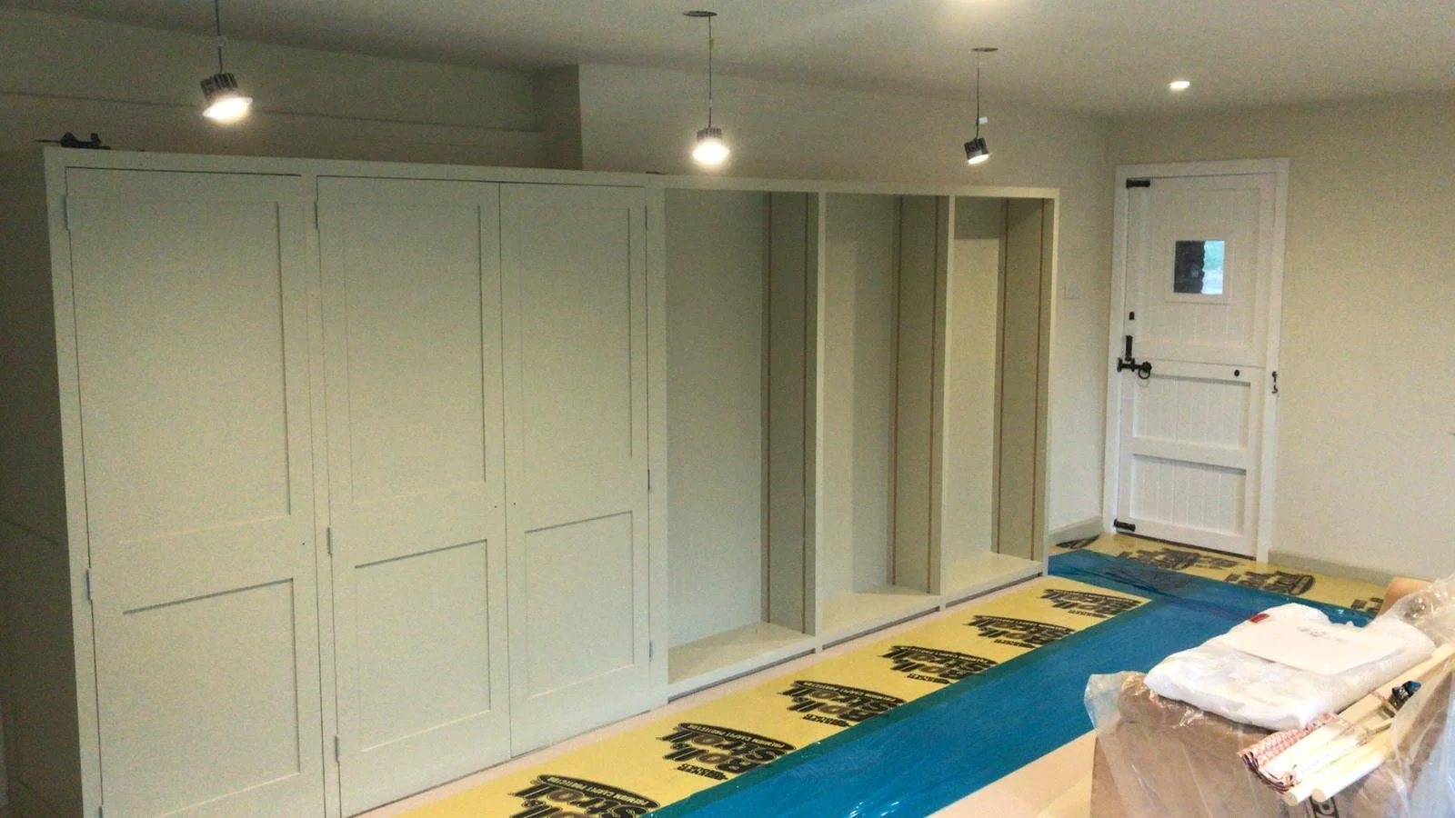 Room under renovation with partially installed white wooden cabinets, protective floor covering, and construction materials.