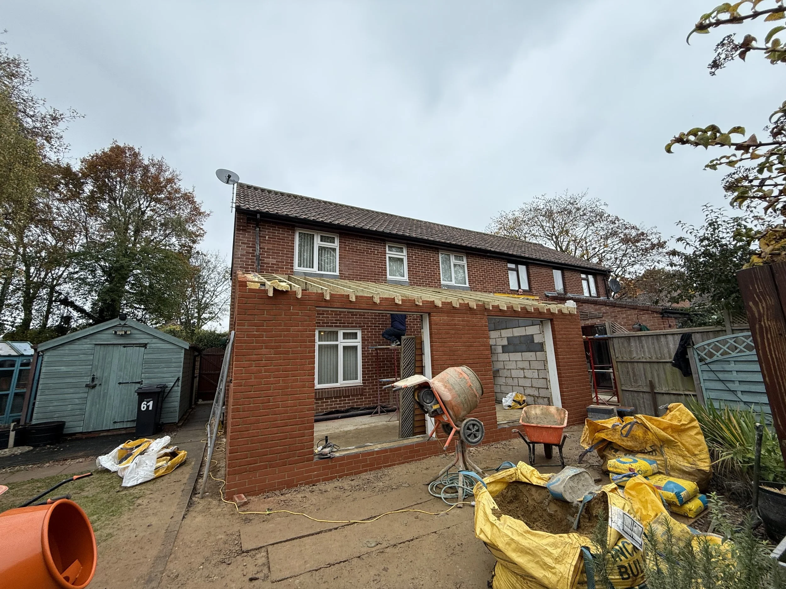 Backyard under construction showing a brick extension being added to a house, with construction tools, cement bags, a wheelbarrow, and scaffolding present.
