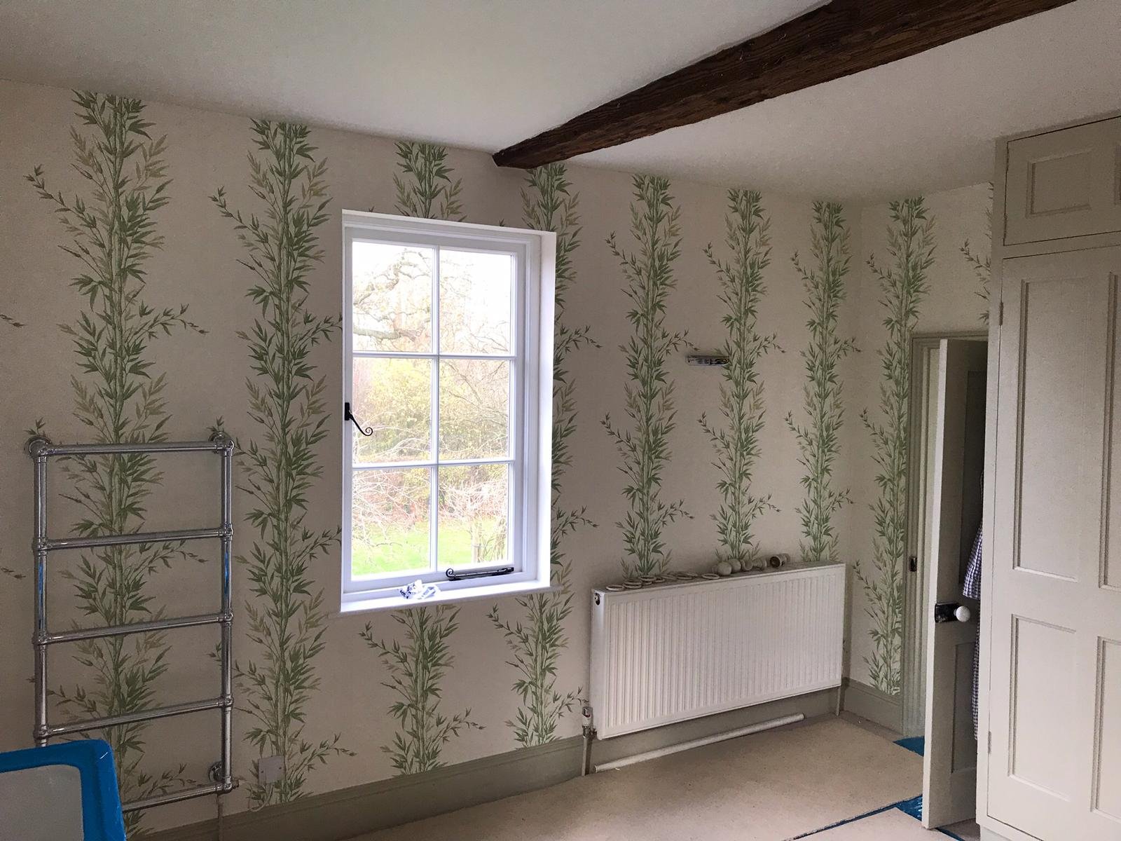 Room with green leafy patterned wallpaper, a window with a view of trees outside, a white radiator, and an unfinished space with a wardrobe and a door.