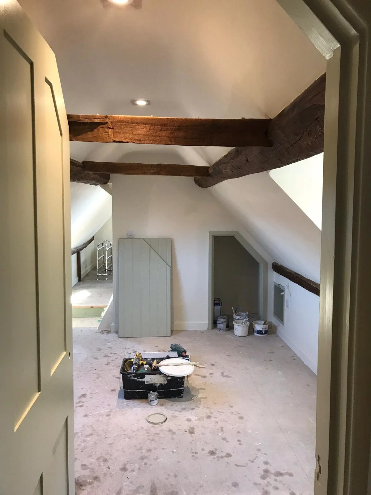 An attic room with sloped ceiling and exposed wooden beams under renovation, with painting and construction tools, paint cans, and a toolbox on the floor.