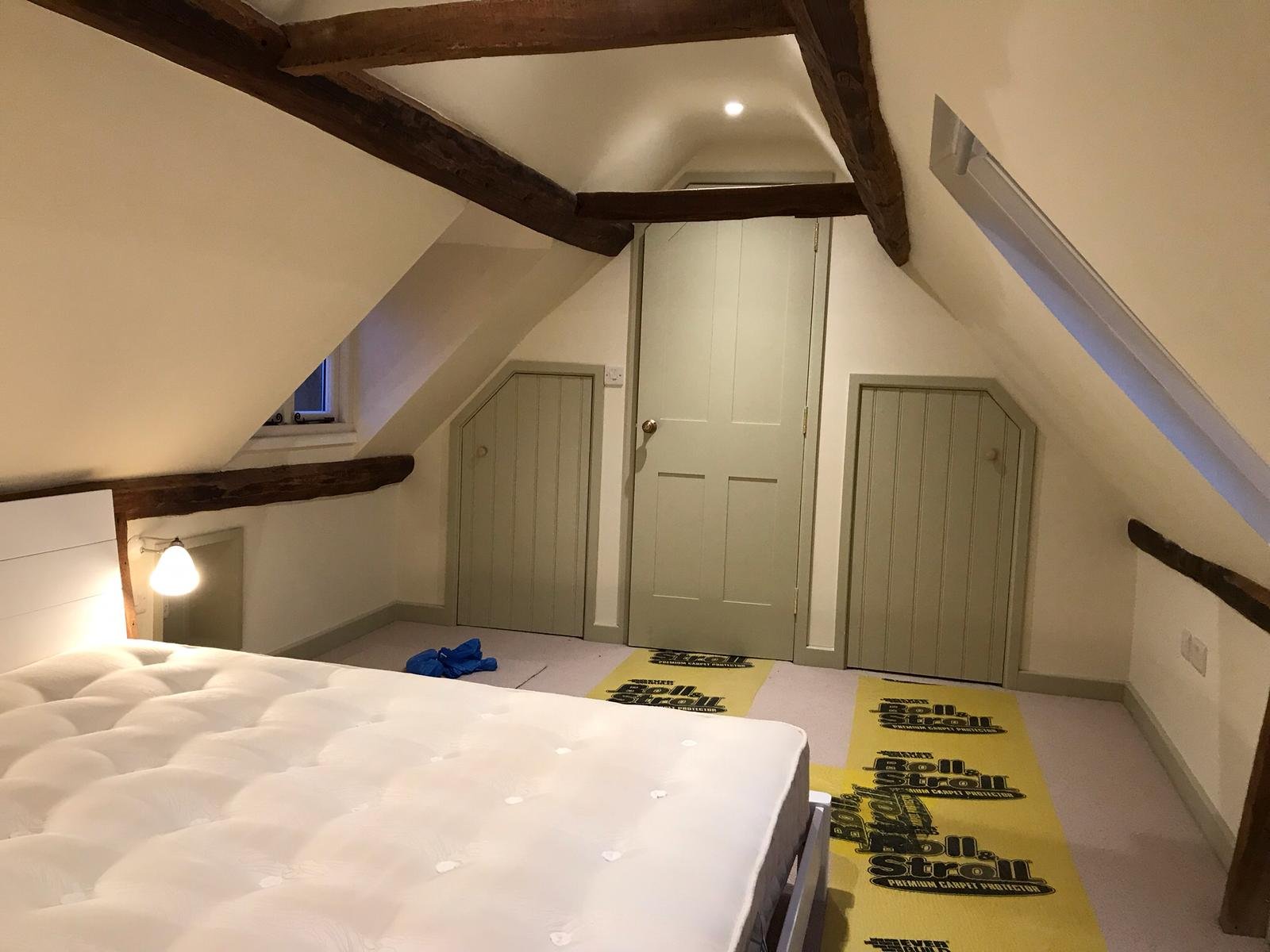 A construction site showing a fenced area with a blue recycling waste container, a grey portable toilet, and a white garage door in the background; and a brick house with a partially installed white bay window and some construction materials nearby.