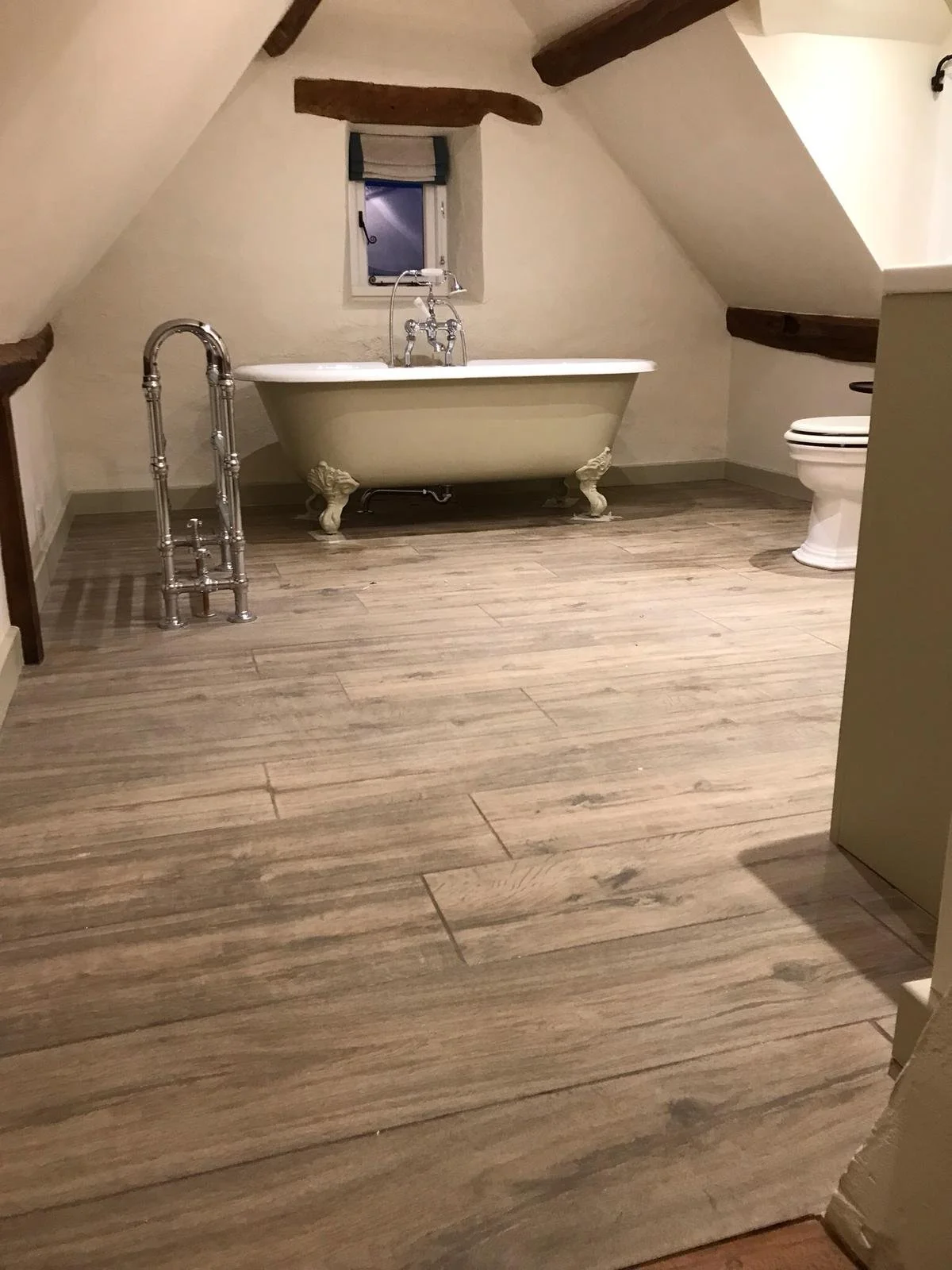 Bathroom with a clawfoot bathtub, toilet, small window, wooden beams on sloped ceiling, and wood-look flooring.