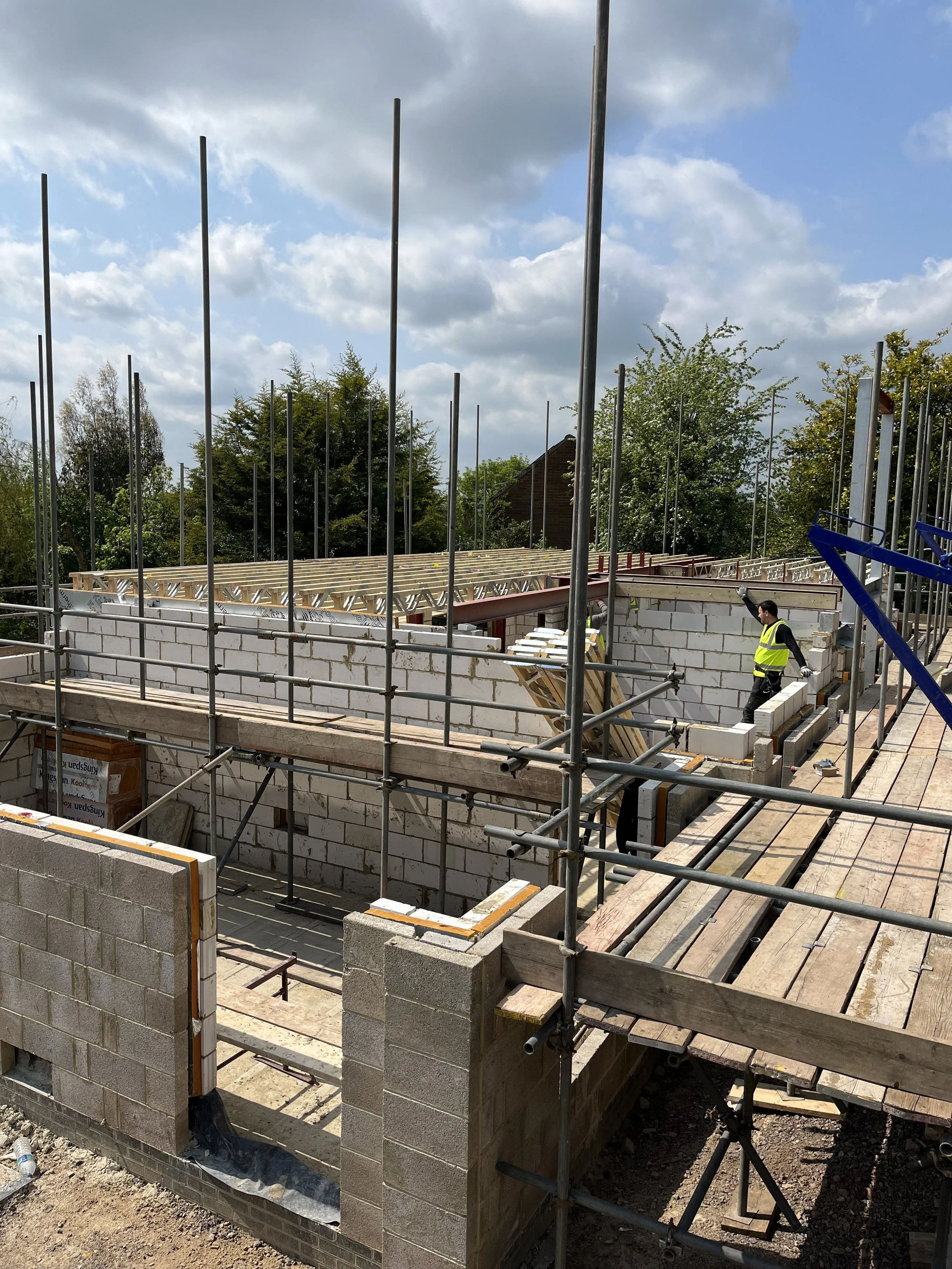 Construction site with foundation framing, construction materials, and workers, under a partly cloudy sky.