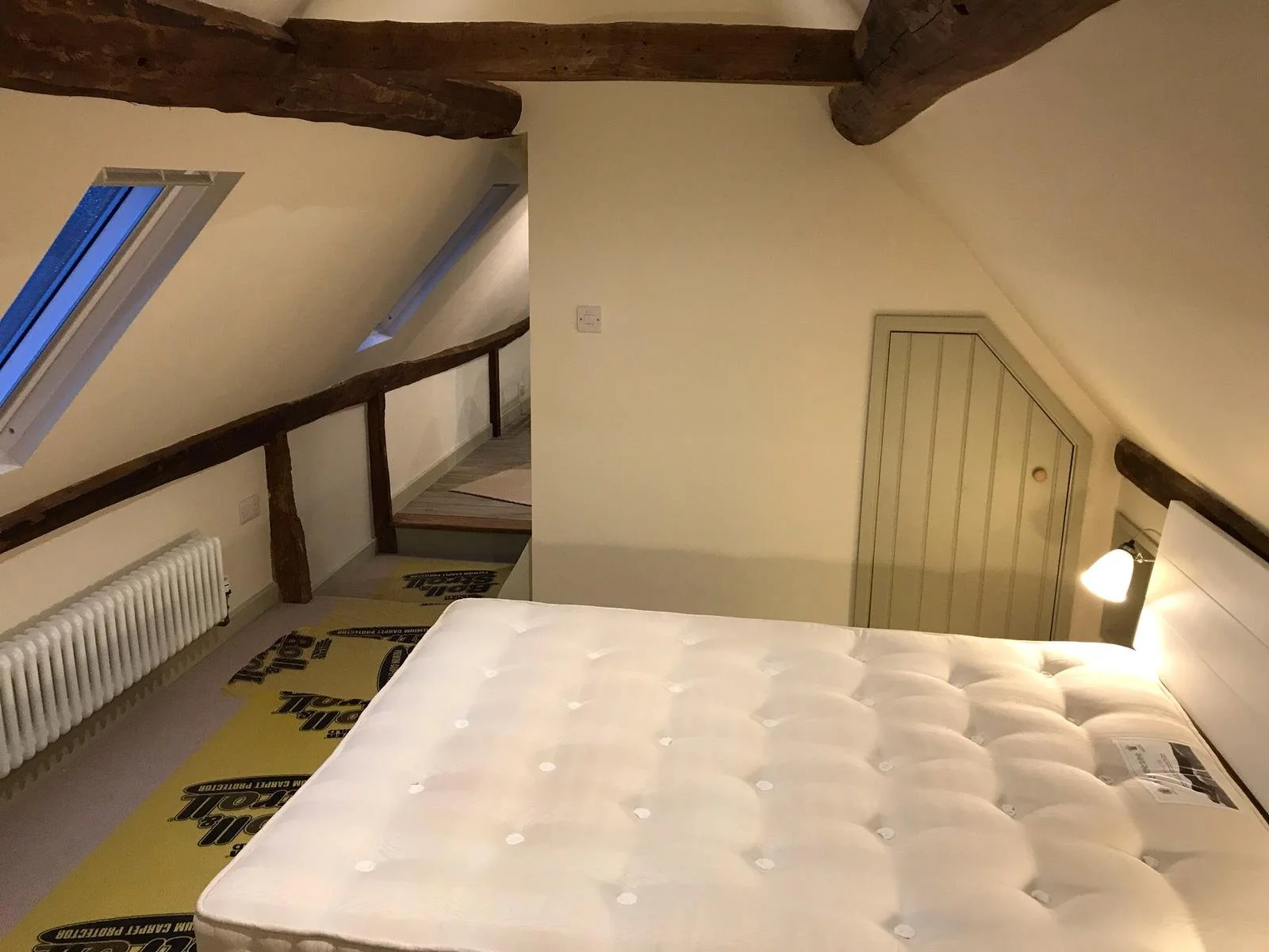 A bedroom with a sloped ceiling, two skylight windows, a white mattress, a white headboard, a wall-mounted light, and wooden beams.