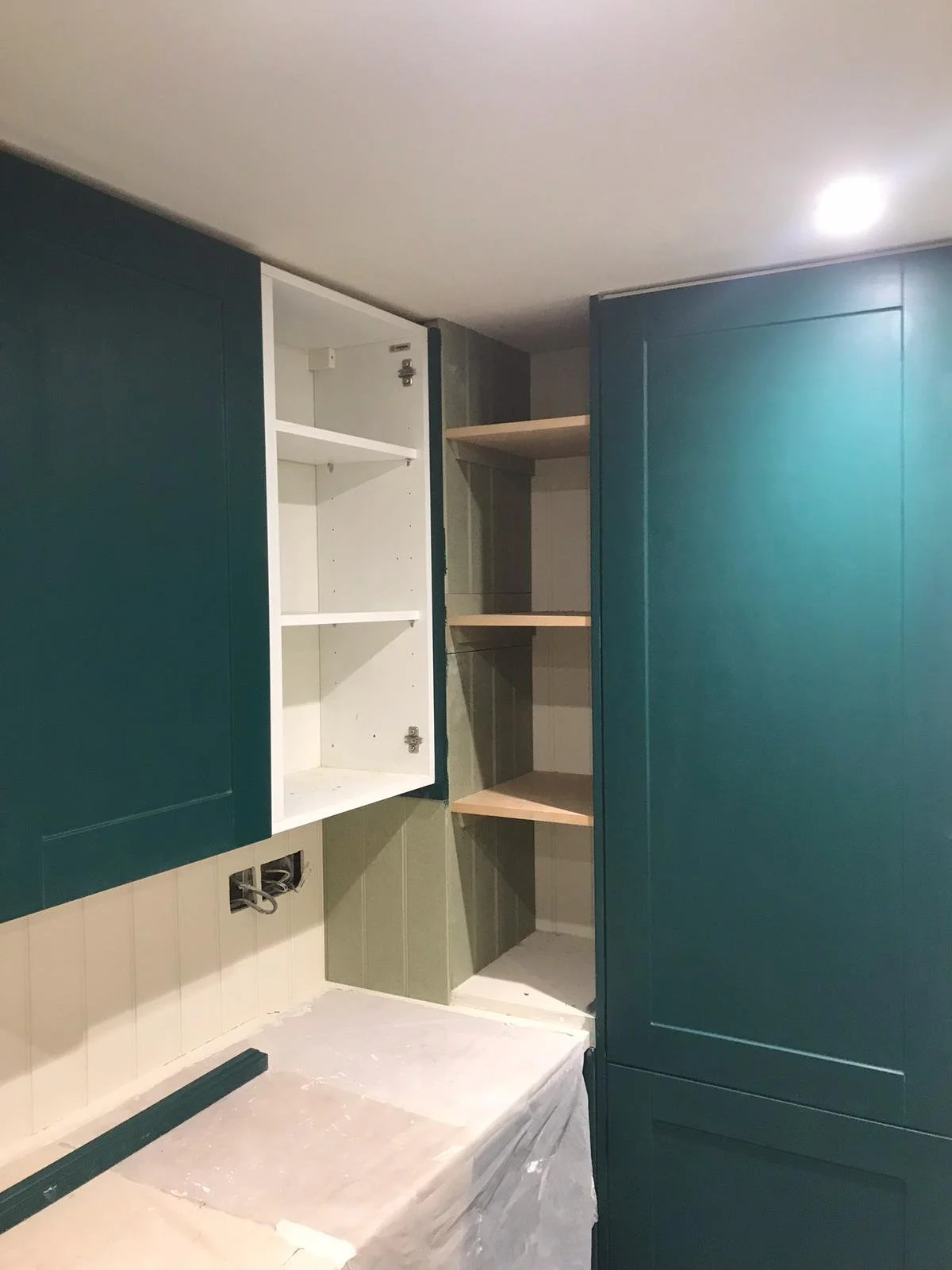 Partially installed kitchen cabinets, with open empty white and beige shelves and dark teal cabinet doors, on a wall with a sink base cabinet and visible electrical wiring.