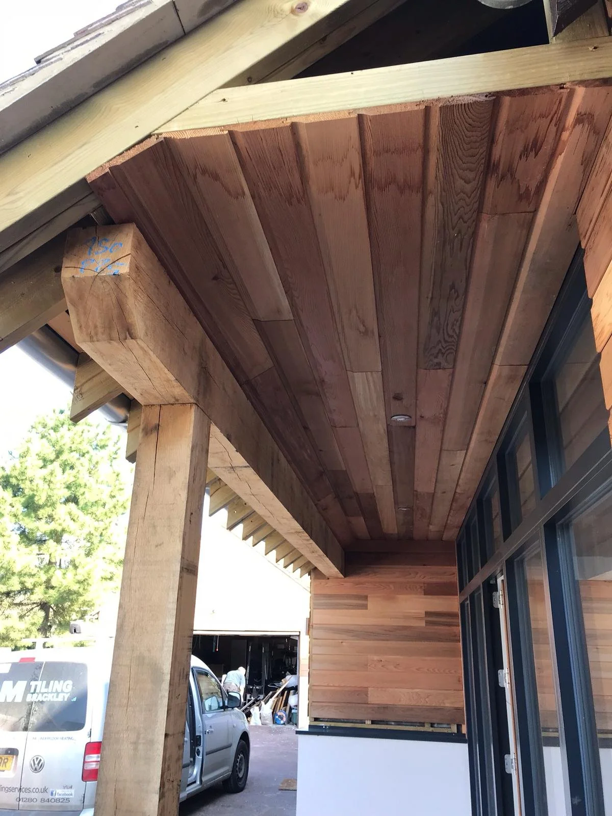 Wooden porch ceiling with a skylight, supported by a vertical wooden post, with a view of a garage and a car in the background.