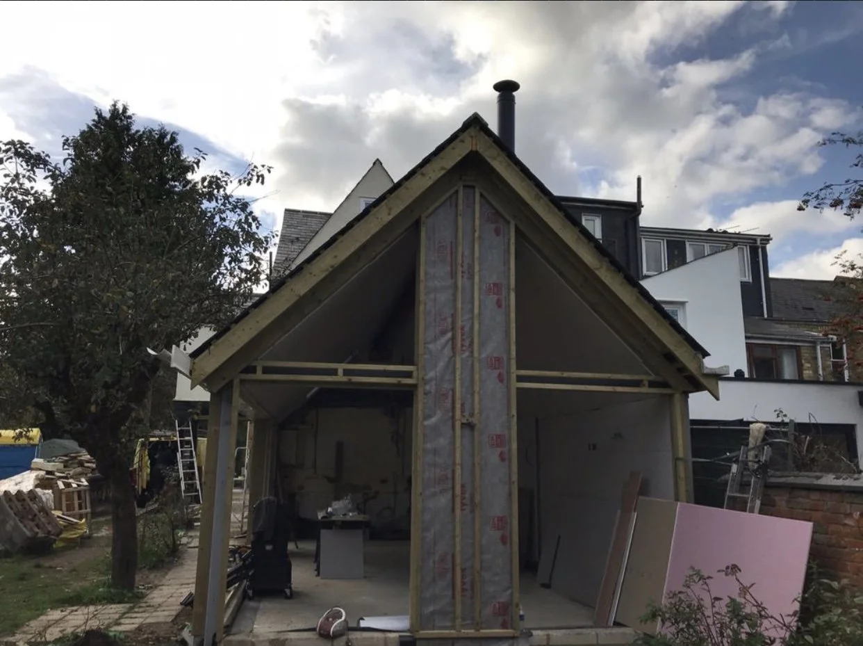 Construction of a small house or extension with wooden framing and insulation, in a residential yard with tools and materials around.