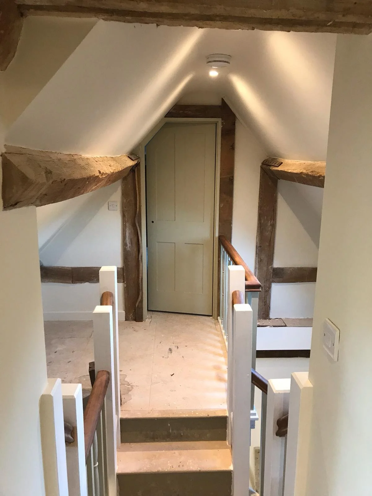 Interior view of a stairway landing with wooden beams, a beige door at the end, and exposed wood framing, with white walls and a wooden handrail.