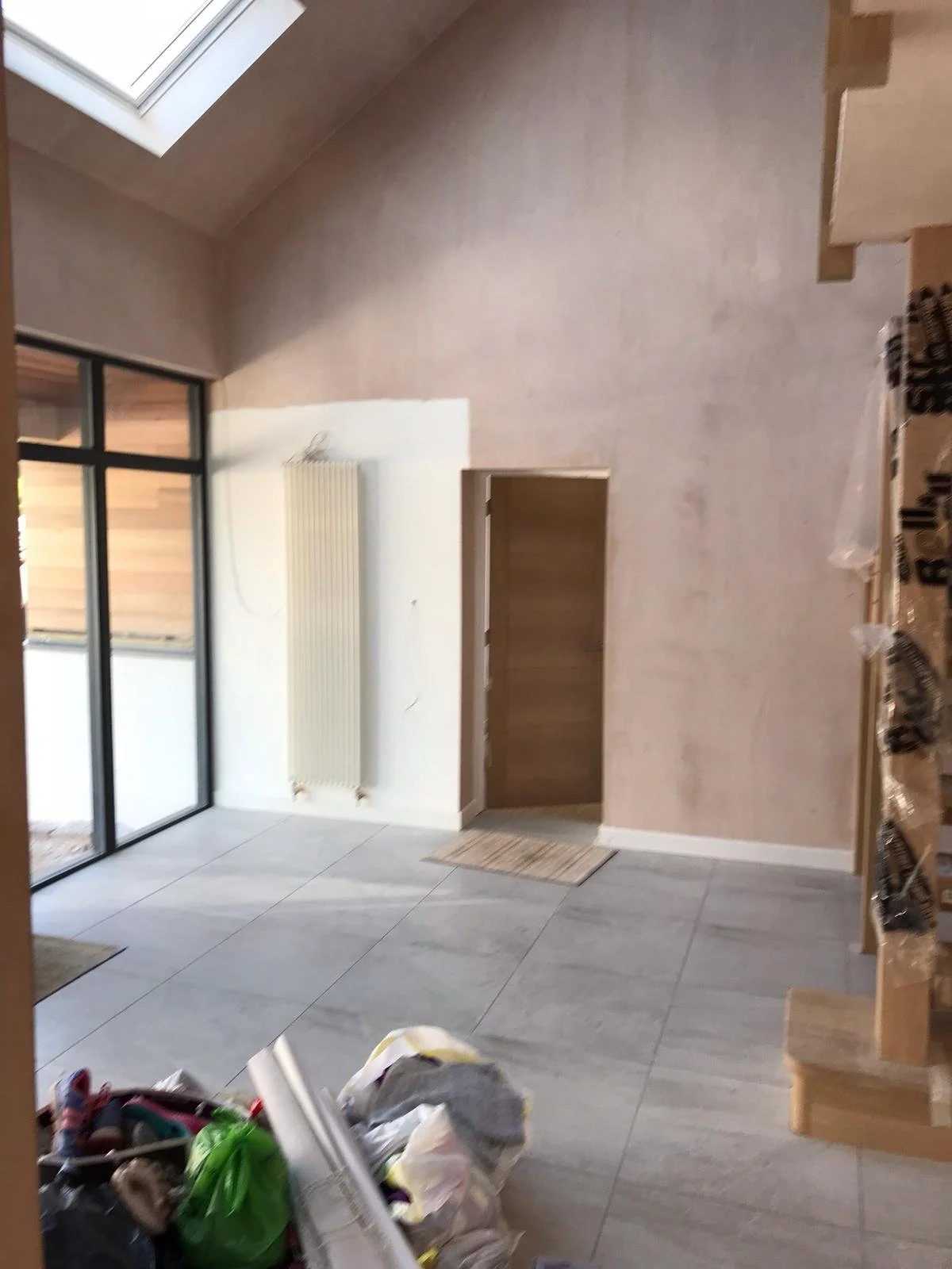 Living room with large window, white radiator, brown door, light-colored tiled floor, and a pile of cleaning supplies or bags in the foreground.