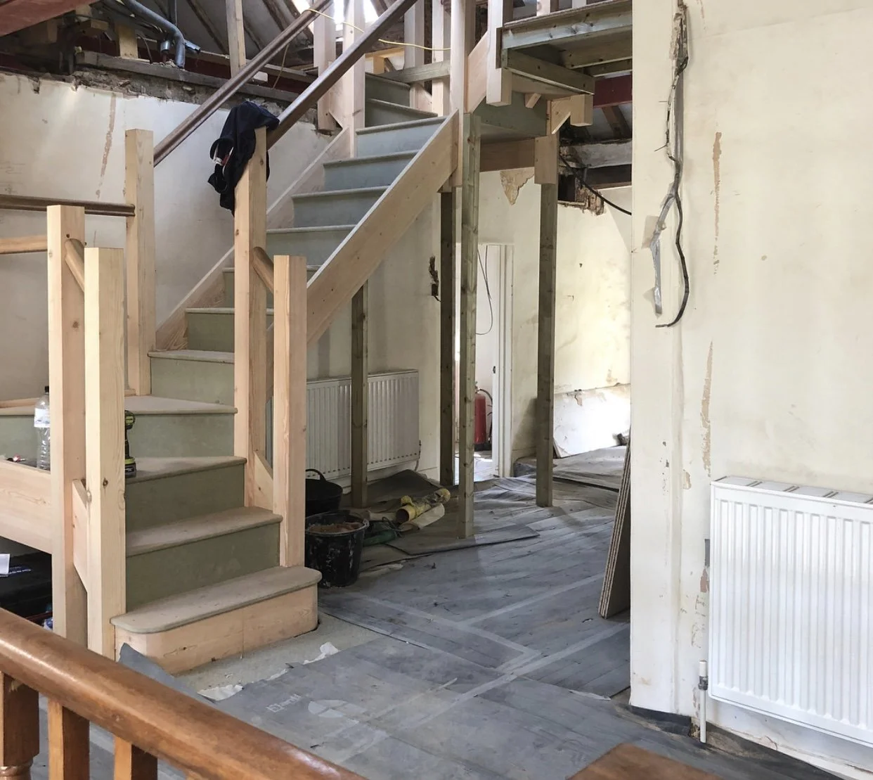 Interior of a house under renovation with a partially built wooden staircase, construction materials, tools, and exposed wiring.