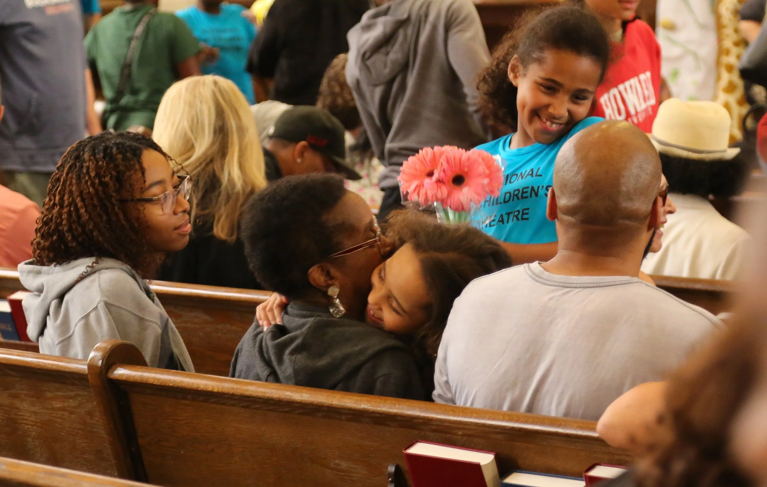 A diverse group of people in a church or community hall, sitting and standing, some engaging with others, with a young girl smiling and holding a pink flower in the foreground.