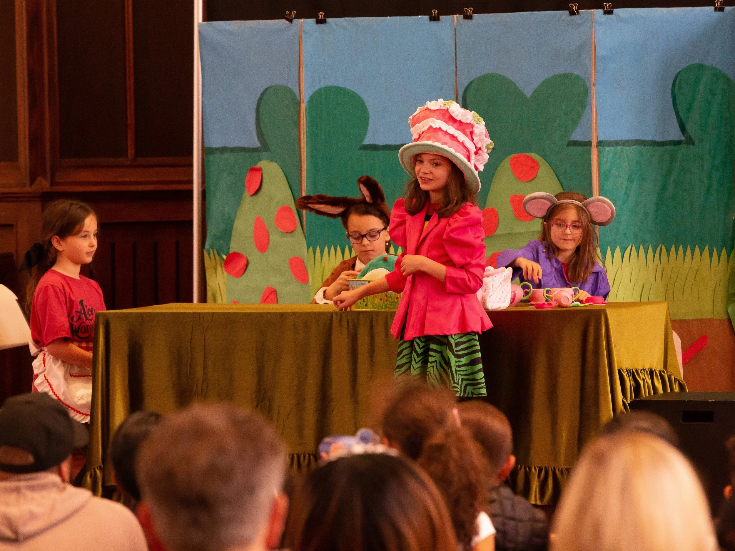 Children performing in a play on stage with a colorful backdrop of trees and hills, some wearing costumes like a large pink hat, bunny ears, and mouse ears.
