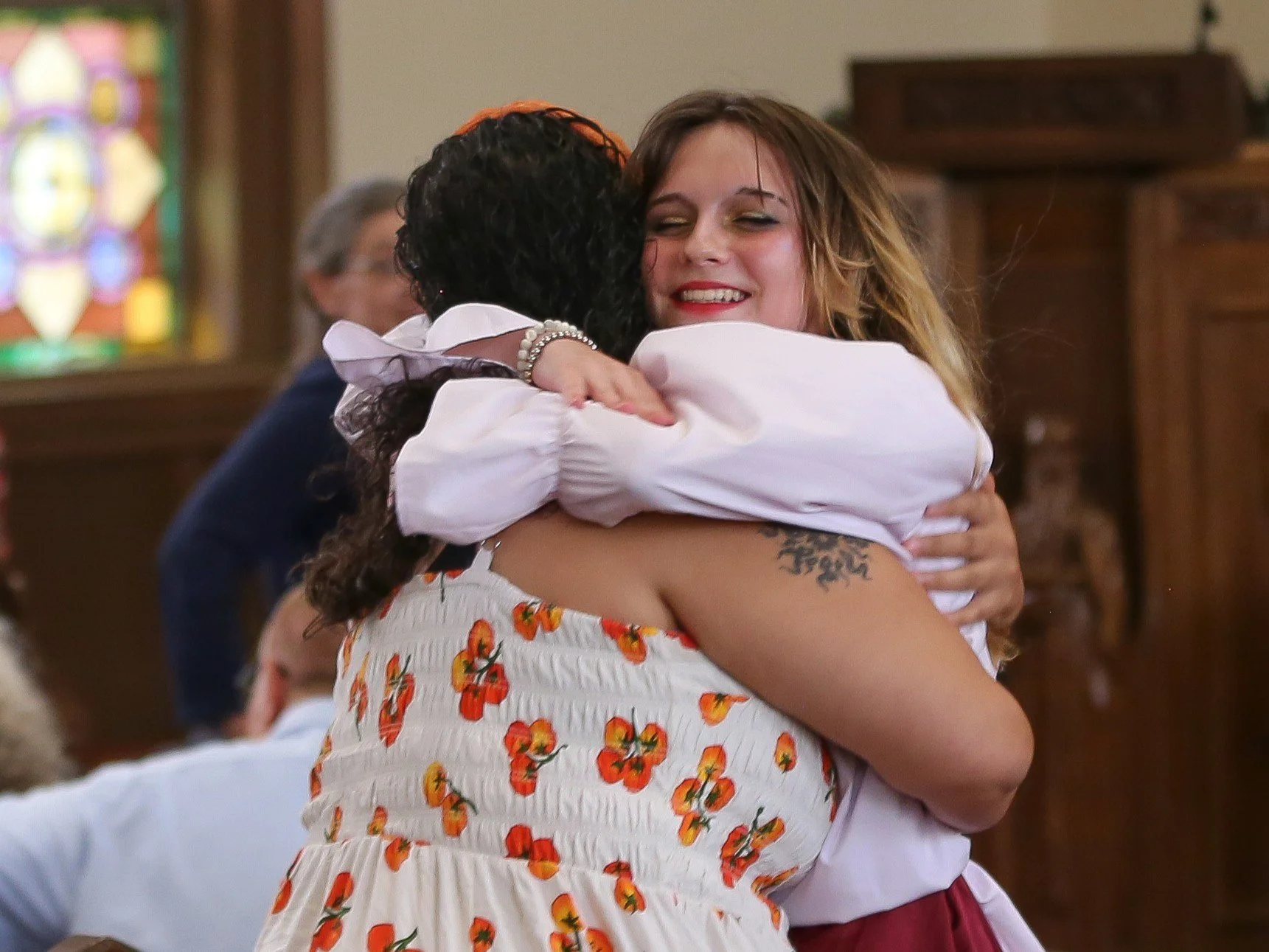 Two women are hugging each other inside a church with stained glass windows. One woman has curly dark hair and is wearing a white dress with an orange floral pattern. The other woman has light brown hair and is wearing a light-colored blouse with gol
