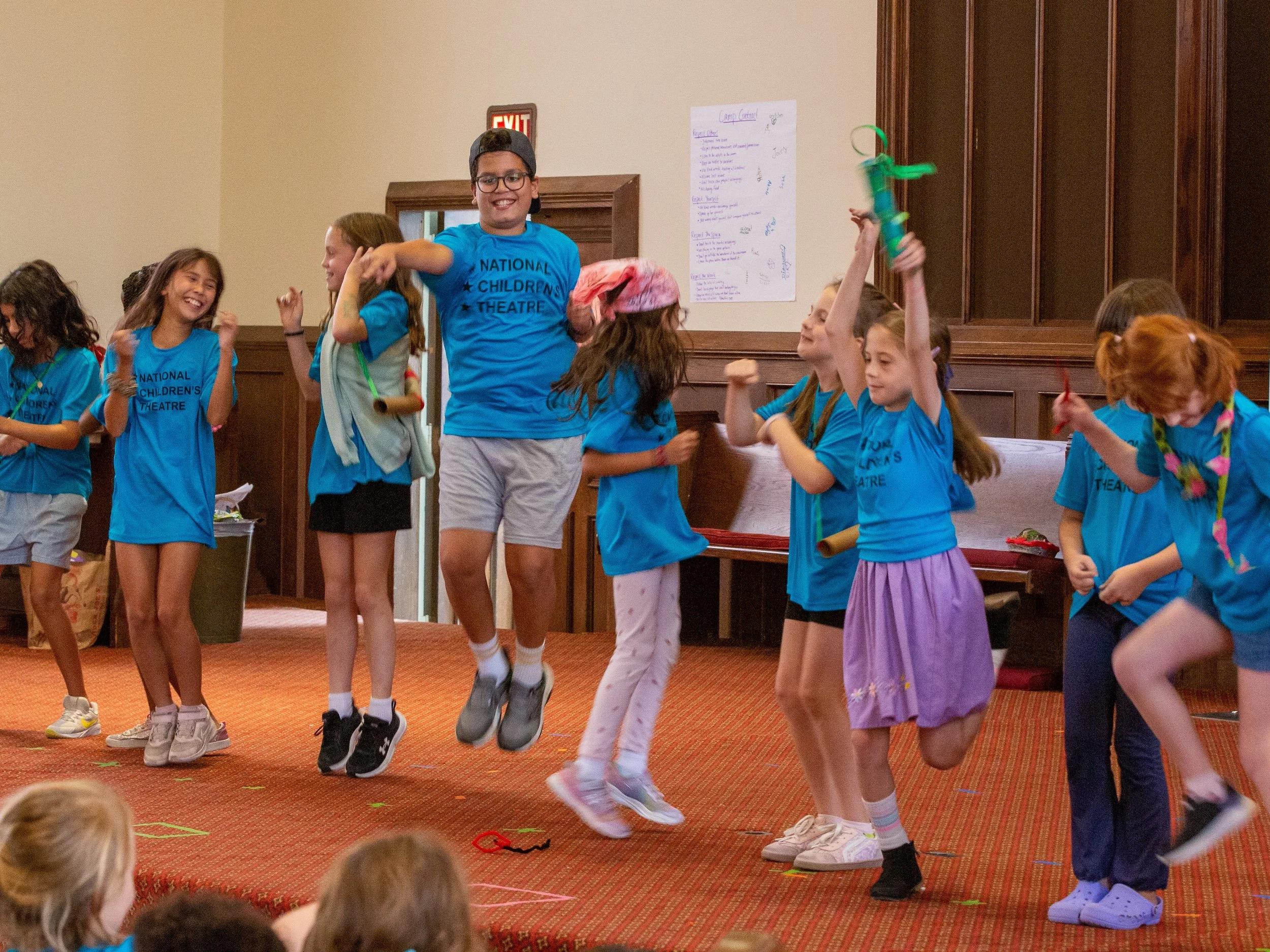 A group of children wearing blue shirts with 'National Children's Theatre' printed on them, actively performing or dancing in an indoor setting, with a boy in a black cap jumping and smiling in the center, surrounded by other children.