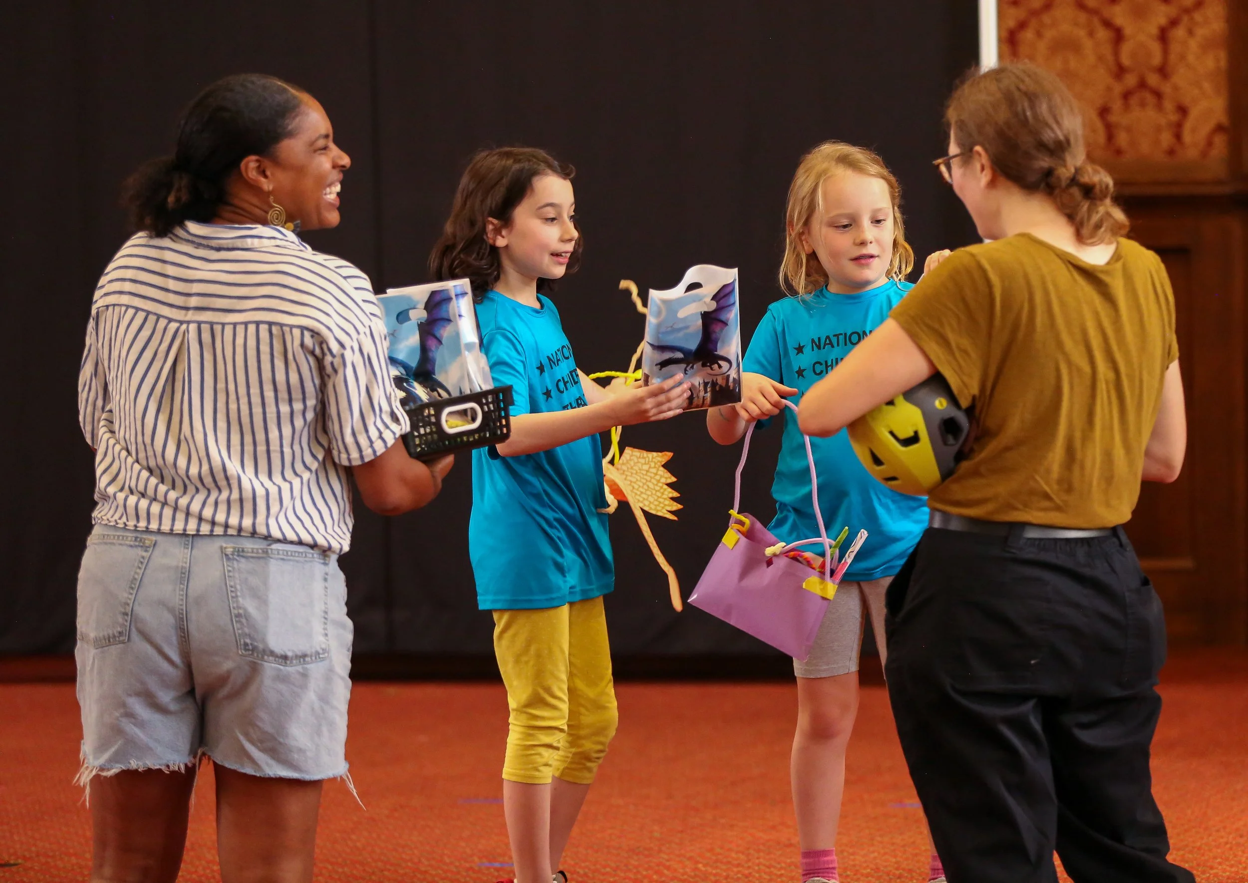 Women and children at an indoor event with a black background. The women are smiling and exchanging gifts or awards, and the children are holding bags with dragon designs.