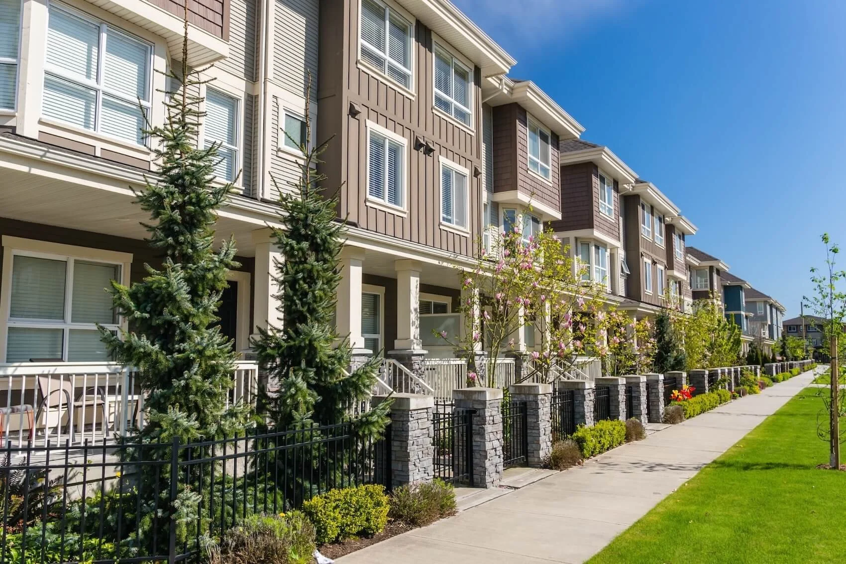A row of modern multi-story townhouses in Shrewsbury, MA with front porches, surrounded by landscaped gardens, trees, and a sidewalk under a clear blue sky.