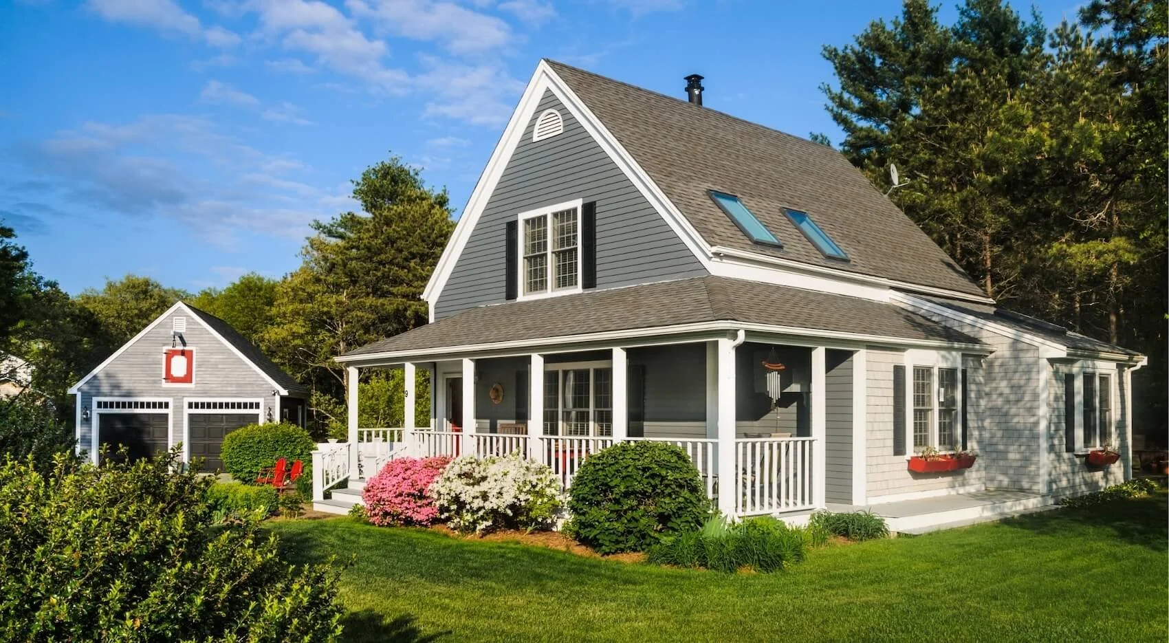 A two-story house with blue siding, white trim, and a dark gray roof, surrounded by a well-maintained garden with colorful flowers and lush green grass, under a bright blue sky.