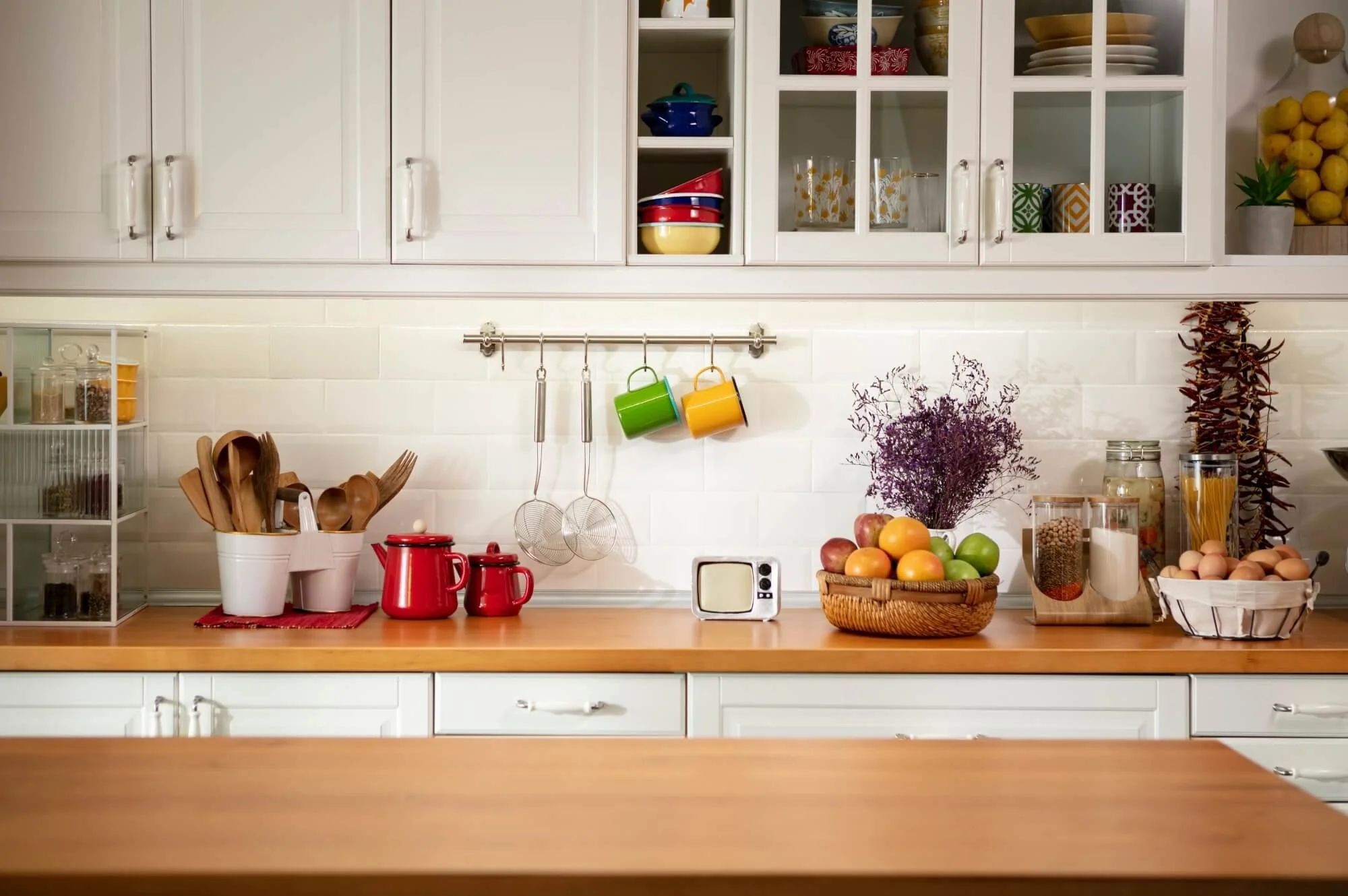 A kitchen countertop with various food and kitchen items, including apples and oranges in a basket, three red teapots, and a container of eggs, and completely free from household pests.