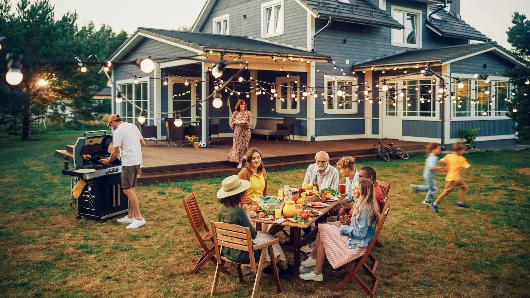 People enjoying an outdoor backyard in Northbridge, MA gathering with string lights, a dining table with food, a barbecue grill, and children running around at sunset.