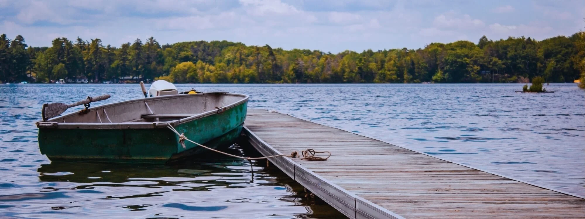 A small green and gray rowboat tied to a wooden dock on a lake in Westborough, MA where shoreline pest control solutions have been applied.