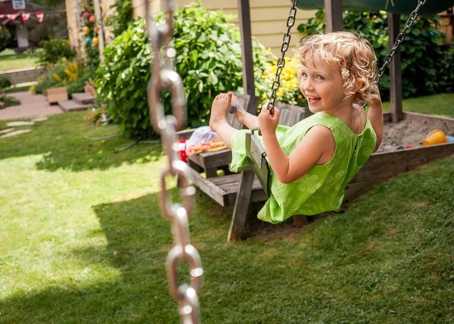 A young girl with curly blonde hair, wearing a green sundress, joyfully swinging on a backyard swing in Millbury, MA.