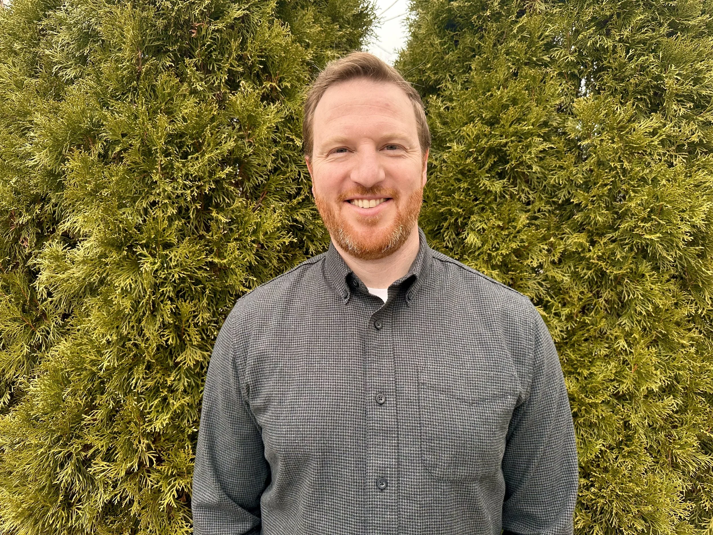 Nick, a person with a beard, short hair, and a checkered shirt, standing against a blue background.