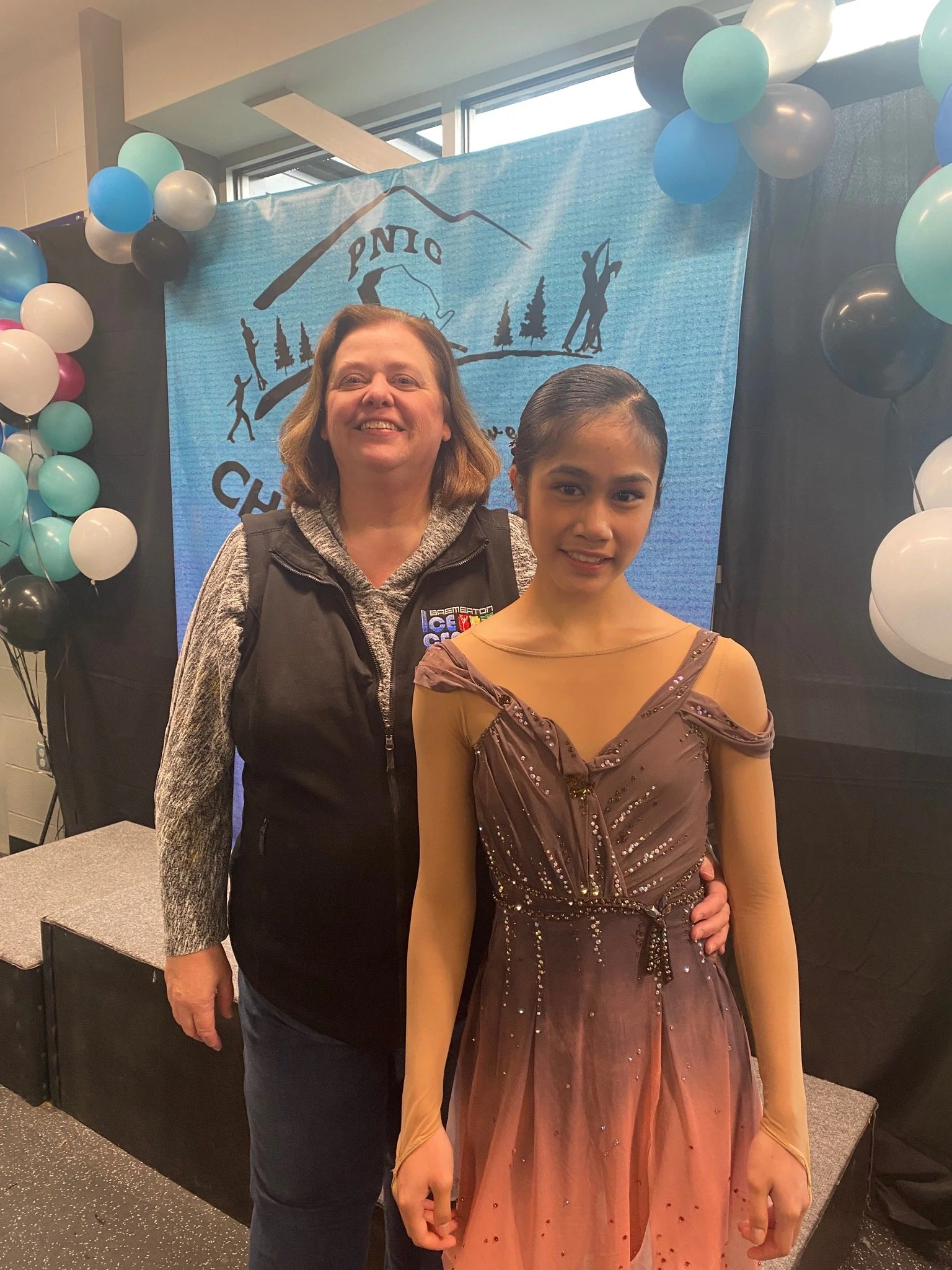 An older woman and a young girl pose together in front of a blue banner with a mountain and pine tree logo, surrounded by colorful balloons at an indoor event.