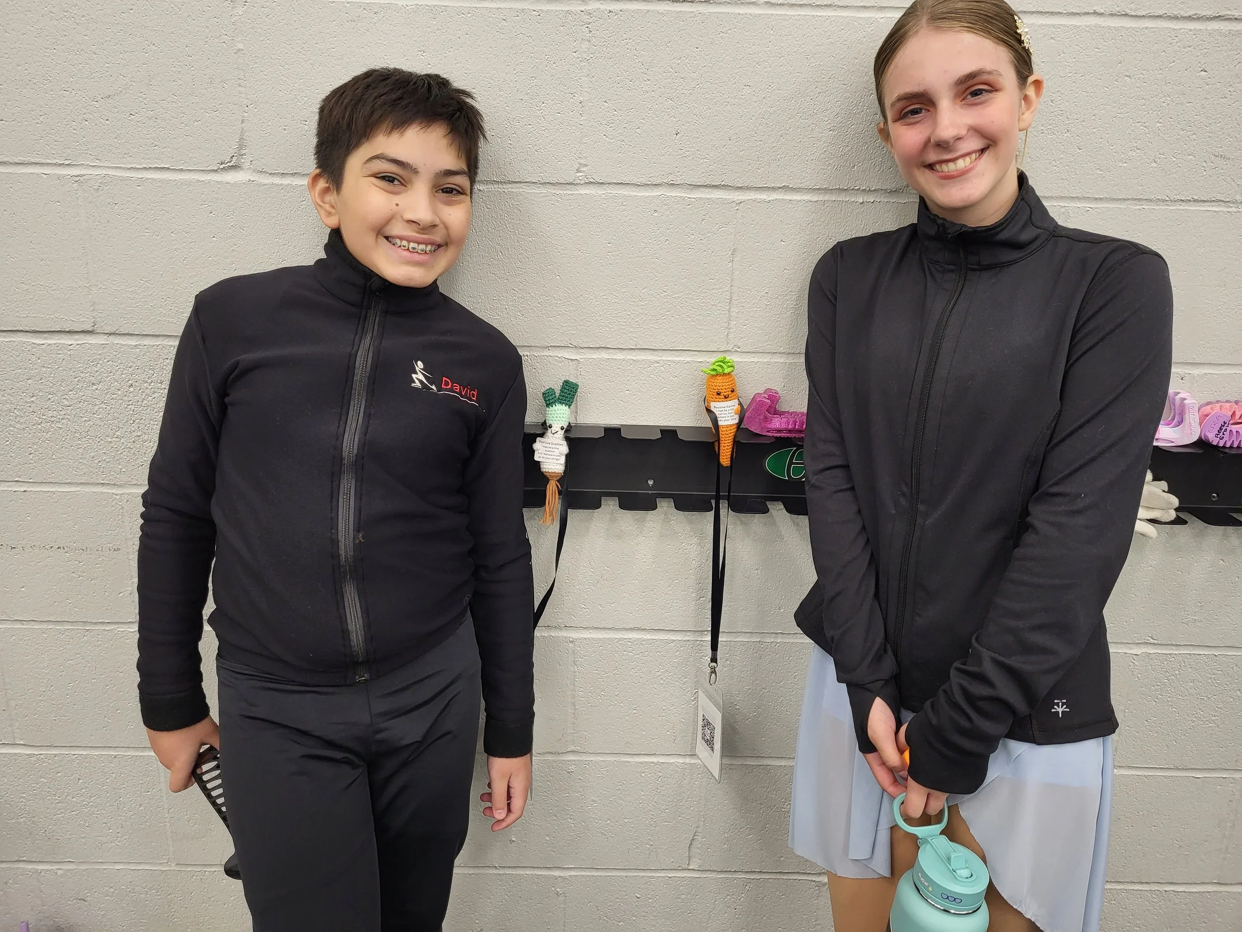 Two young girls smiling and standing against a cinder block wall with hooks holding handmade knitted vegetable decorations and pink slippers.